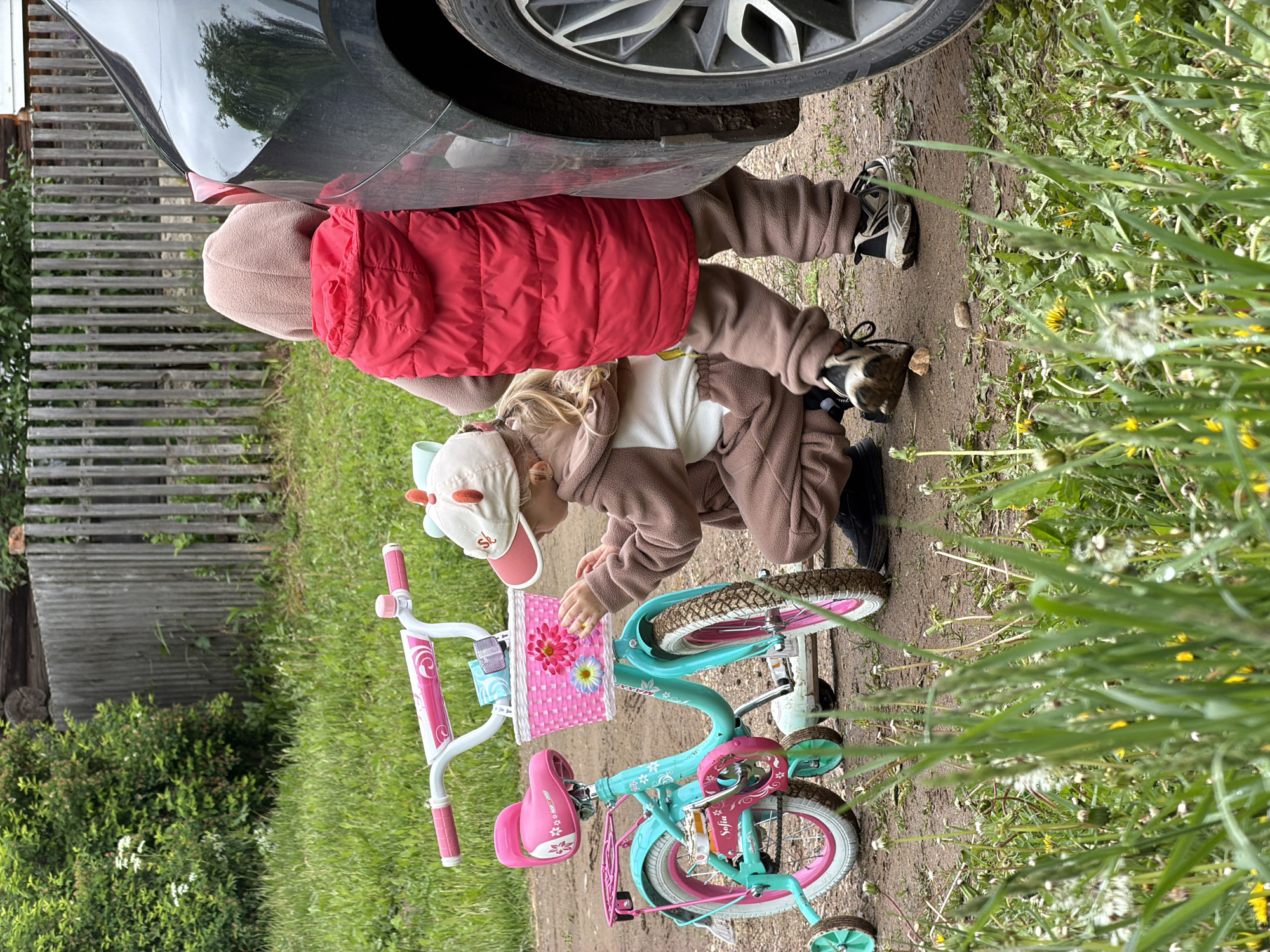 The image captures an outdoor scene featuring two young children, a car, and a bicycle, set in what appears to be a rural or semi-rural environment in Marakushi, Russia. The perspective is tilted, showing the ground and subjects vertically.

One child, seen mostly from the back, wears a bright red puffer jacket over a brown fleece outfit and dark sneakers. This child stands close to the rear wheel of a dark-colored car, whose shiny surface reflects surrounding greenery. The word "OUTSIDE" is partially visible on the car's tire.

The second child, dressed in a brown fleece jumpsuit and a light-colored cap with what resemble animal ears and an "S" emblem, is crouching next to a child's bicycle. The bicycle is turquoise with pink accents on the seat, handlebars, and training wheels. A pink woven basket, adorned with a pink and a yellow/blue flower, is attached to the handlebars. The child's hands are near the basket, suggesting interaction with it.

The scene takes place on a dirt path, bordered by lush green grass and various plants, including dandelions in different stages of bloom. In the background, a rustic wooden fence stretches horizontally, with bushes and trees visible beyond it. The overall impression is a calm, daytime activity in a natural setting.