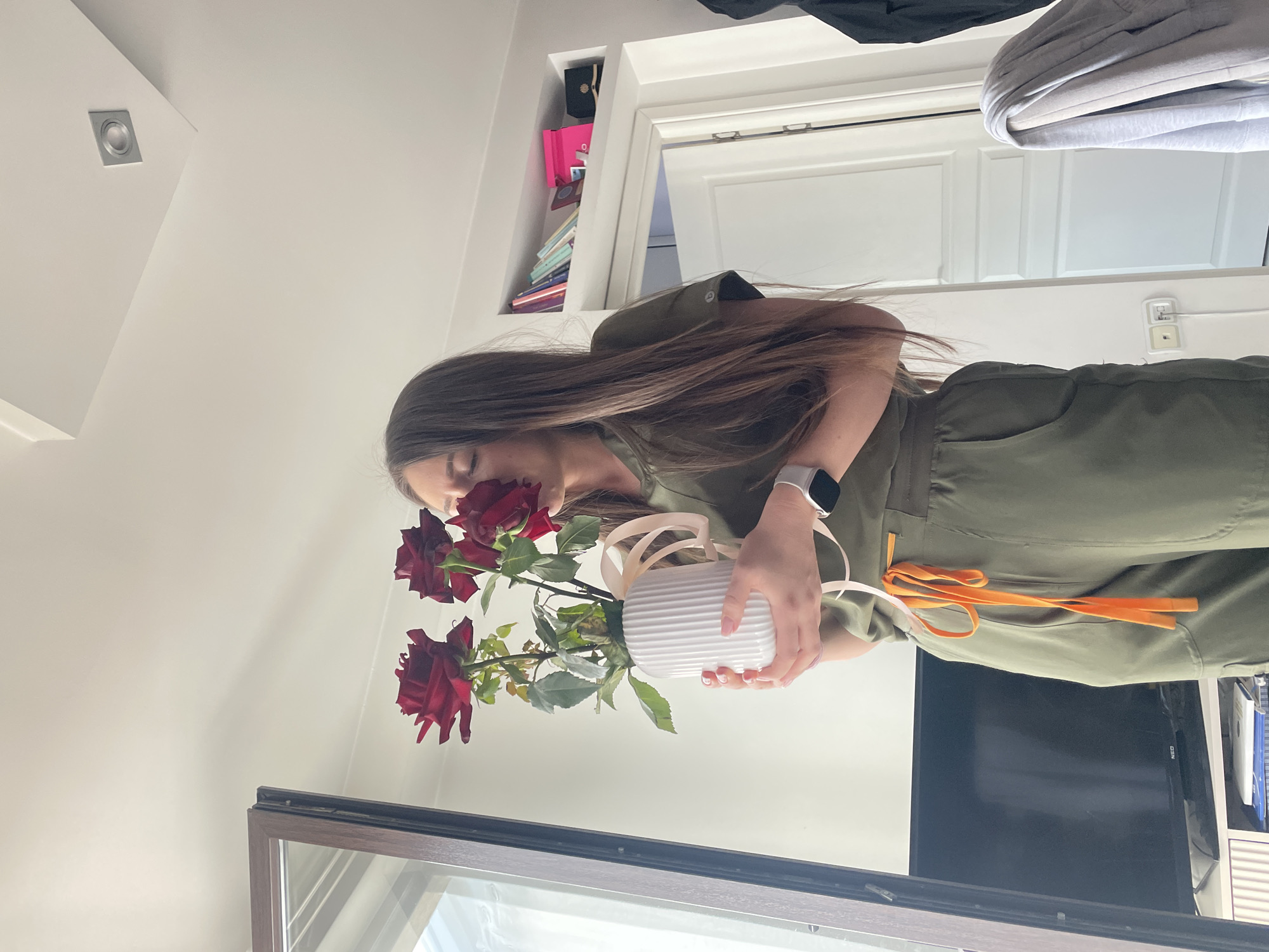 A young woman with long, brown hair is holding a white vase with red roses in an indoor setting. She is wearing an olive-green jumpsuit and a white smartwatch. The vase is decorated with a beige ribbon. The background includes a white wall with a light fixture, a doorway with shelves above, and a TV. There's a reflection visible in a window frame at the bottom of the image. The location cannot be identified as Sofia, Bulgaria, without additional information.