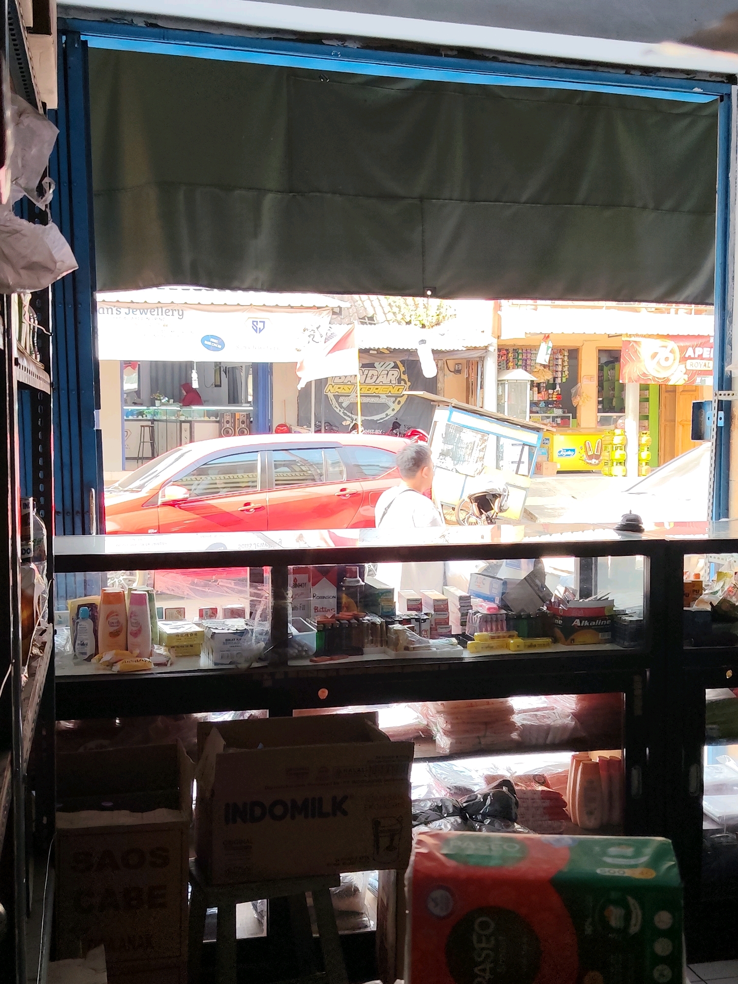The image depicts the interior of a small shop, possibly a convenience store or warung, viewed from behind a counter. In the foreground, a glass display case contains various items, including bottles of lotion or shampoo, boxes, and other products. Behind the counter, shelves are stocked with more goods. Two cardboard boxes are visible, one labeled "SAOS CABE" (chili sauce) and another "INDOMILK". To the right, a package of "PASEO" tissues is prominently displayed.
Through an open doorway, the street scene outside is visible. A red car is parked on the street, and people are seen inside the shop. Buildings and other street elements suggest an urban environment. The overall impression is that of a busy local business operating in Dampit, Indonesia.