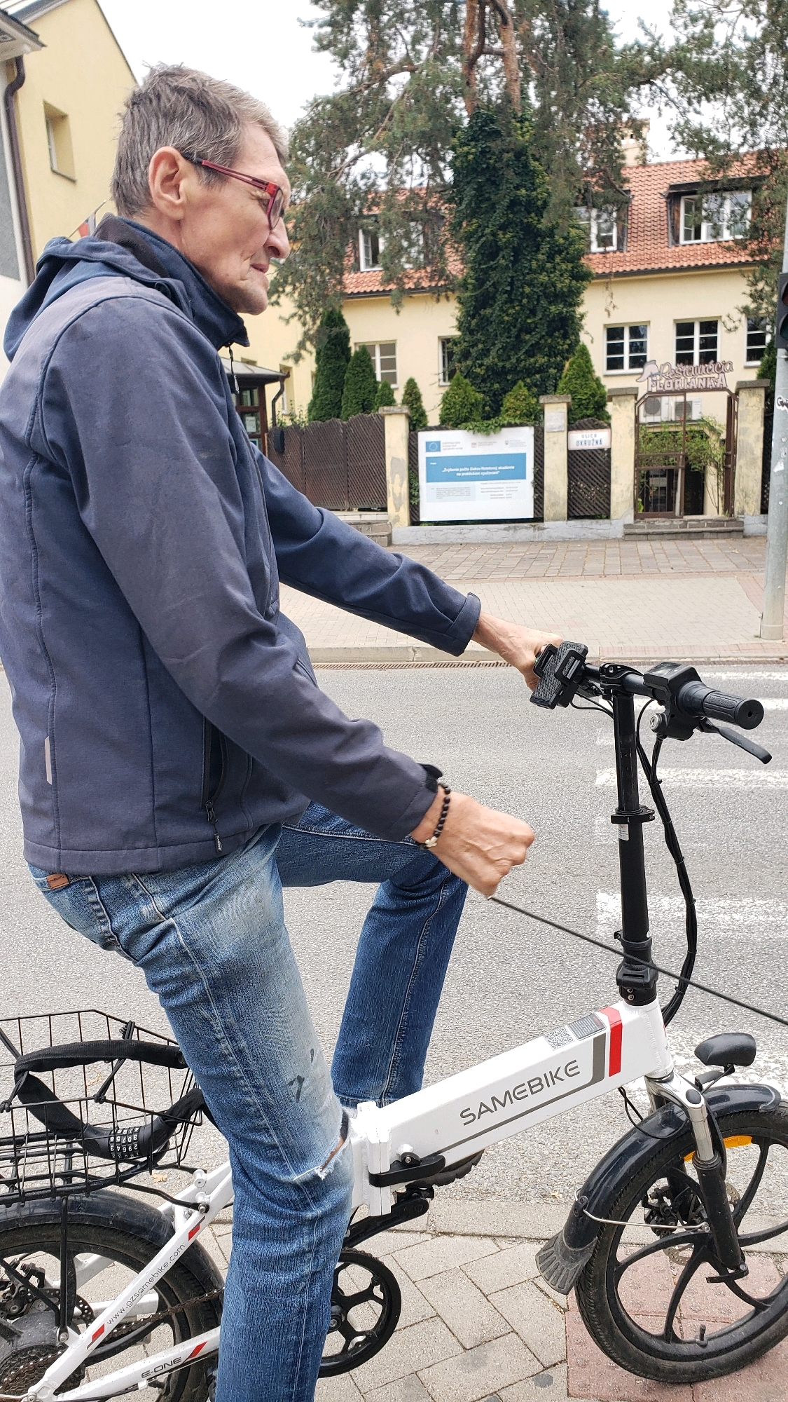 A middle-aged man with glasses and gray hair is riding a white foldable electric bicycle with "SAMEBIKE" branding. He is wearing a dark blue jacket and blue jeans, with a black bicycle lock attached to the rear rack. The man is on a street in the city of Prešov, Slovakia, with buildings and trees in the background. One building has a sign that reads "Reštaurácia U Gringu." The scene appears to be outdoors on a paved street.
