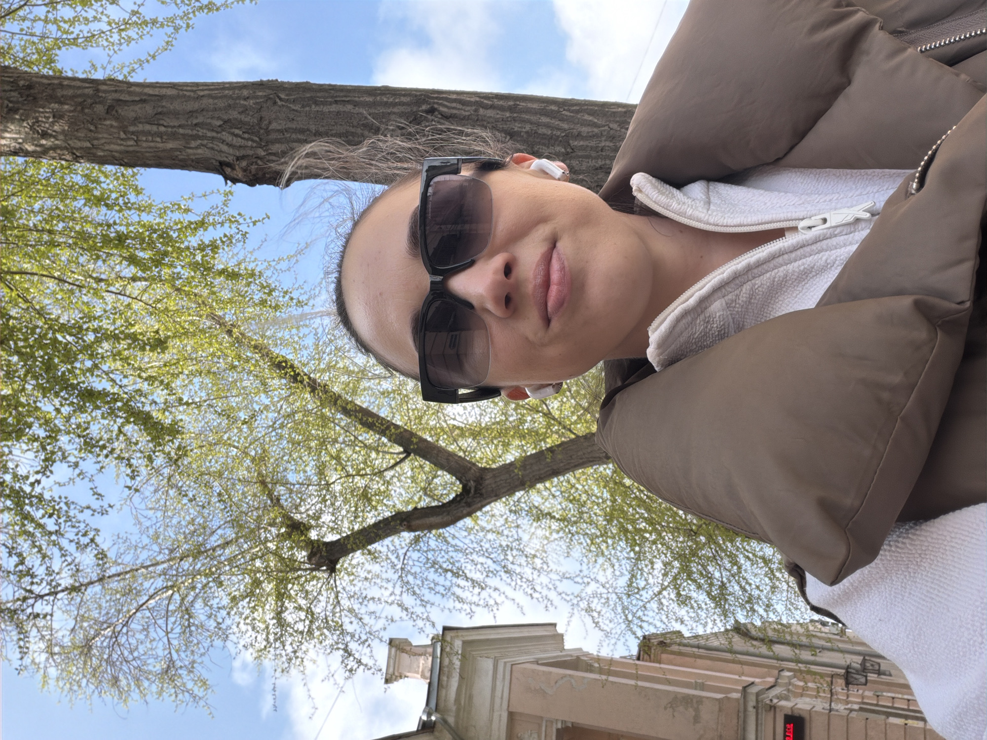 A selfie of a woman with sunglasses, a white zippered jacket, and a brown puffer coat, likely in Rostov-on-Don, Russia. The background includes tree branches with green leaves and building facades.