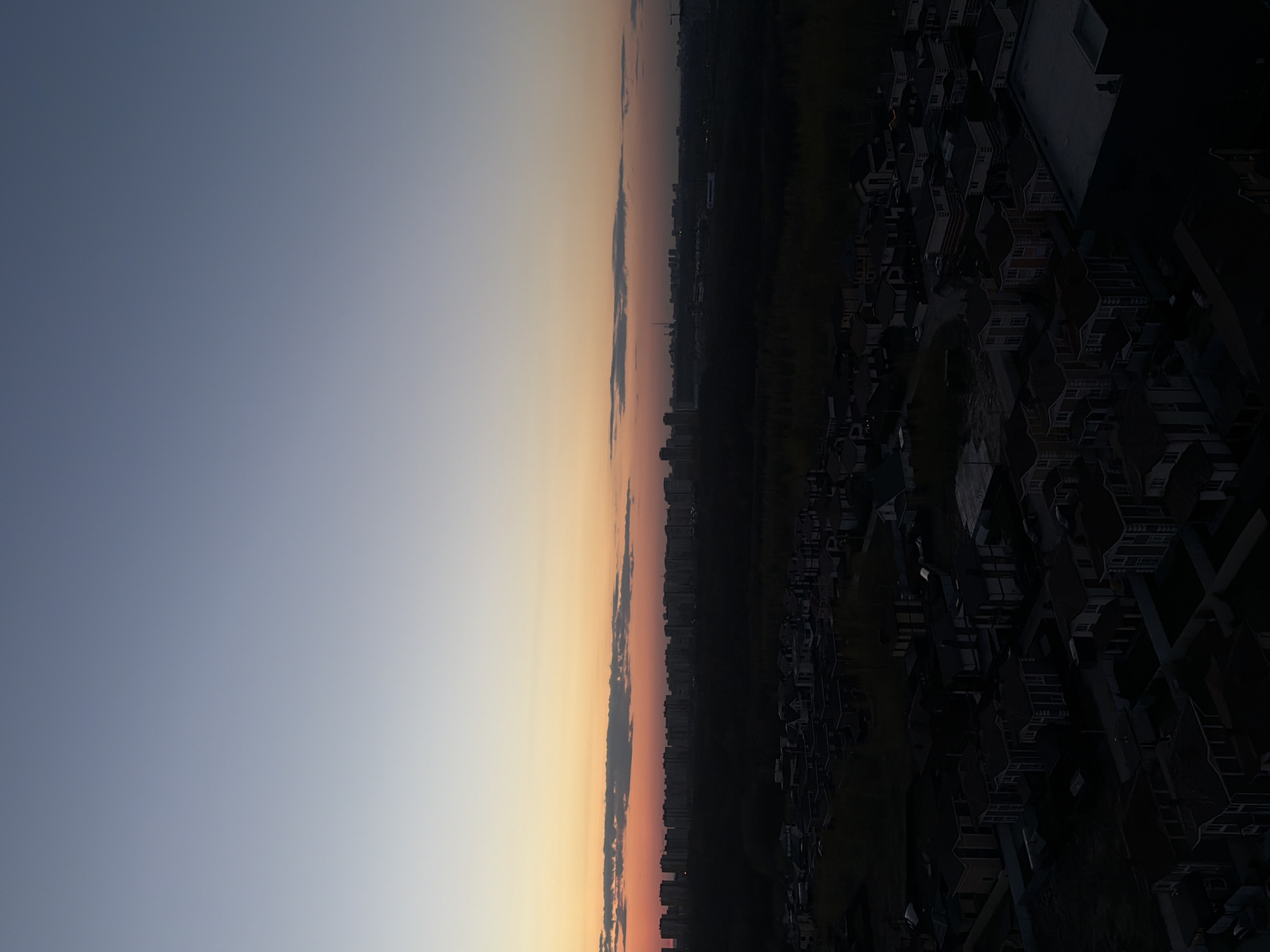 The image shows an aerial view of Анкудиновка, Russia at sunset or sunrise. The sky transitions from blue to orange. A line of clouds is visible, and beyond it, the skyline of a distant cityscape. The foreground shows residential buildings and green space. The buildings appear dark and silhouetted.