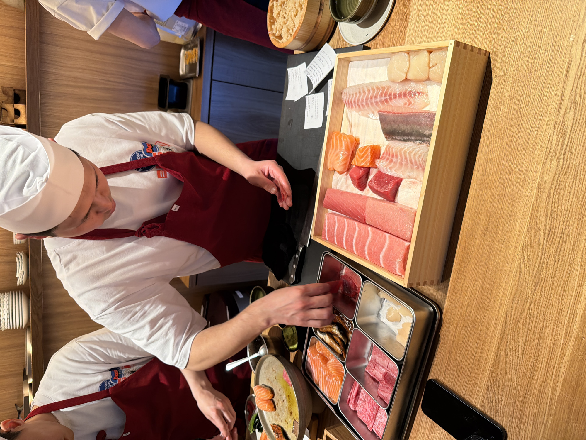 This image, captured in Moscow, Russia, depicts a vibrant scene of sushi and sashimi preparation at a professional counter. Two chefs, wearing white chef's hats, white t-shirts, and red aprons, are diligently working. The chef in the foreground is actively selecting ingredients from a compartmentalized metal tray with his right hand, while his left hand rests near a circular platter holding prepared sushi, including salmon nigiri.
The counter, made of light-colored wood with a noticeable grain, is laden with fresh ingredients. A large wooden box showcases an array of meticulously arranged raw fish fillets and scallops, including various cuts of tuna, salmon, and white fish. The metal tray holds additional selections, appearing to include more tuna, salmon, possibly marbled beef or very fatty fish, and eel. A small wooden bowl contains what looks like grated ginger or another condiment.
In the background, another chef is partially visible, mirroring the attire of the first. The setting includes stacked white dishes, small green bowls, and several white order slips or receipts taped to a dark panel behind the chefs. A small black electronic device, possibly a smartphone, rests on the wooden counter. The lighting is bright, suggesting daytime, and illuminates the food, emphasizing its freshness. On the primary chef's white t-shirt, a circular logo is partially visible, featuring text that appears to include "TON" and "Day." The scene conveys a sense of focused culinary artistry in a Japanese restaurant or sushi bar environment.