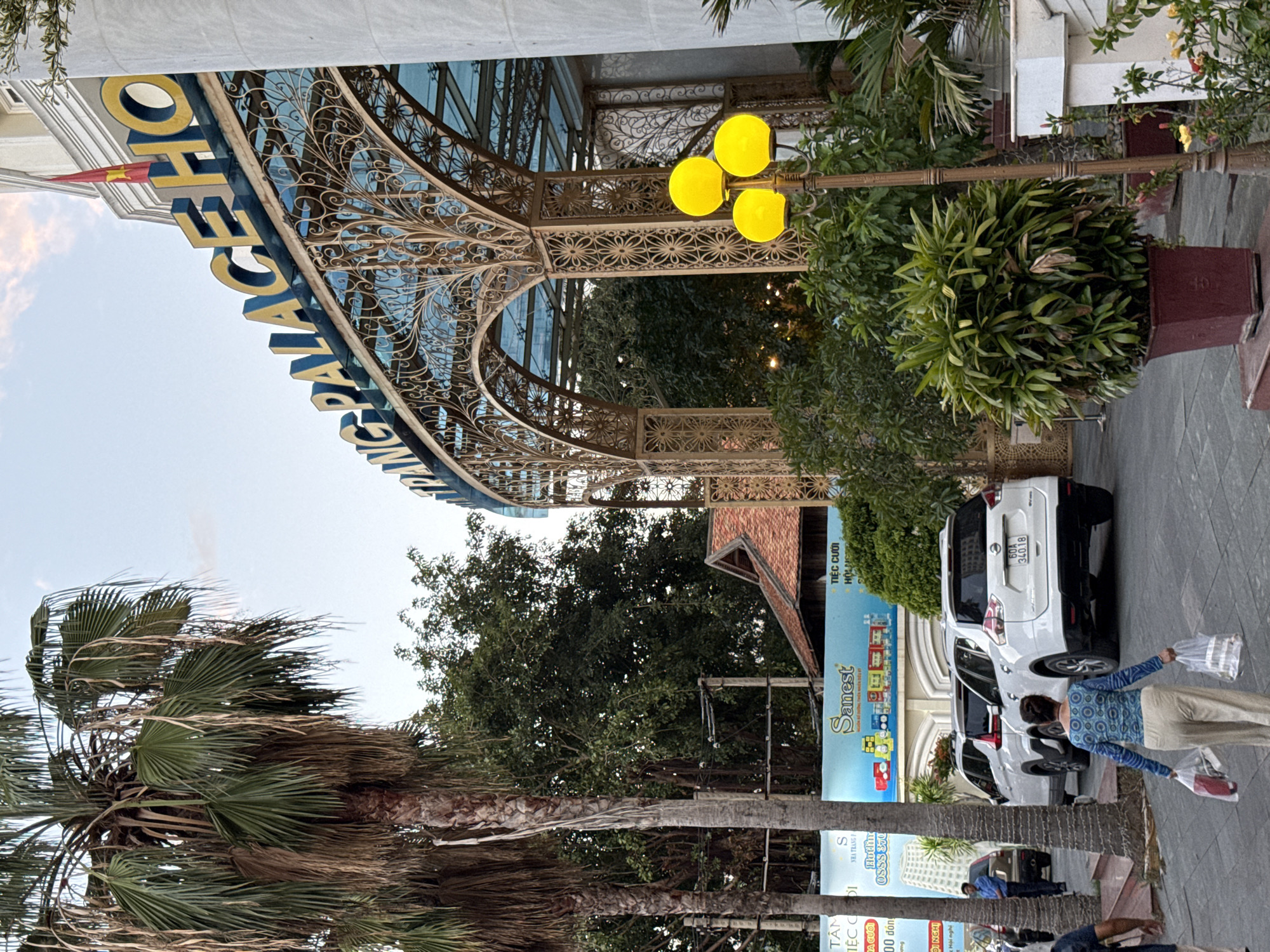 The image depicts the entrance to the "TRANG PALACE HOTEL" in Nha Trang, Vietnam. The hotel's name is prominently displayed in large yellow letters with blue outlines against a blue sky. The entrance features an ornate golden metal archway with intricate floral patterns, leading to a covered walkway with a glass roof. Tall palm trees are visible to the left of the hotel entrance, with their fronds reaching upwards.
In the foreground, a woman walks away from the viewer, carrying two plastic shopping bags in each hand. She is wearing a light-colored long-sleeved shirt with a blue patterned design and light-colored pants. Behind her, two white SUVs are parked. The license plate of the leading SUV reads "60A 34018". A security guard in a blue uniform stands near the entrance.
A large blue billboard is visible on the left side of the image, featuring text and images, including the word "Sanest". The environment appears to be a street or sidewalk in front of the hotel, with paved ground and some greenery. A decorative lamp post with three yellow spherical lights stands to the right of the entrance, surrounded by dense green foliage in a dark red planter. The overall scene suggests a sunny day in a tropical city.