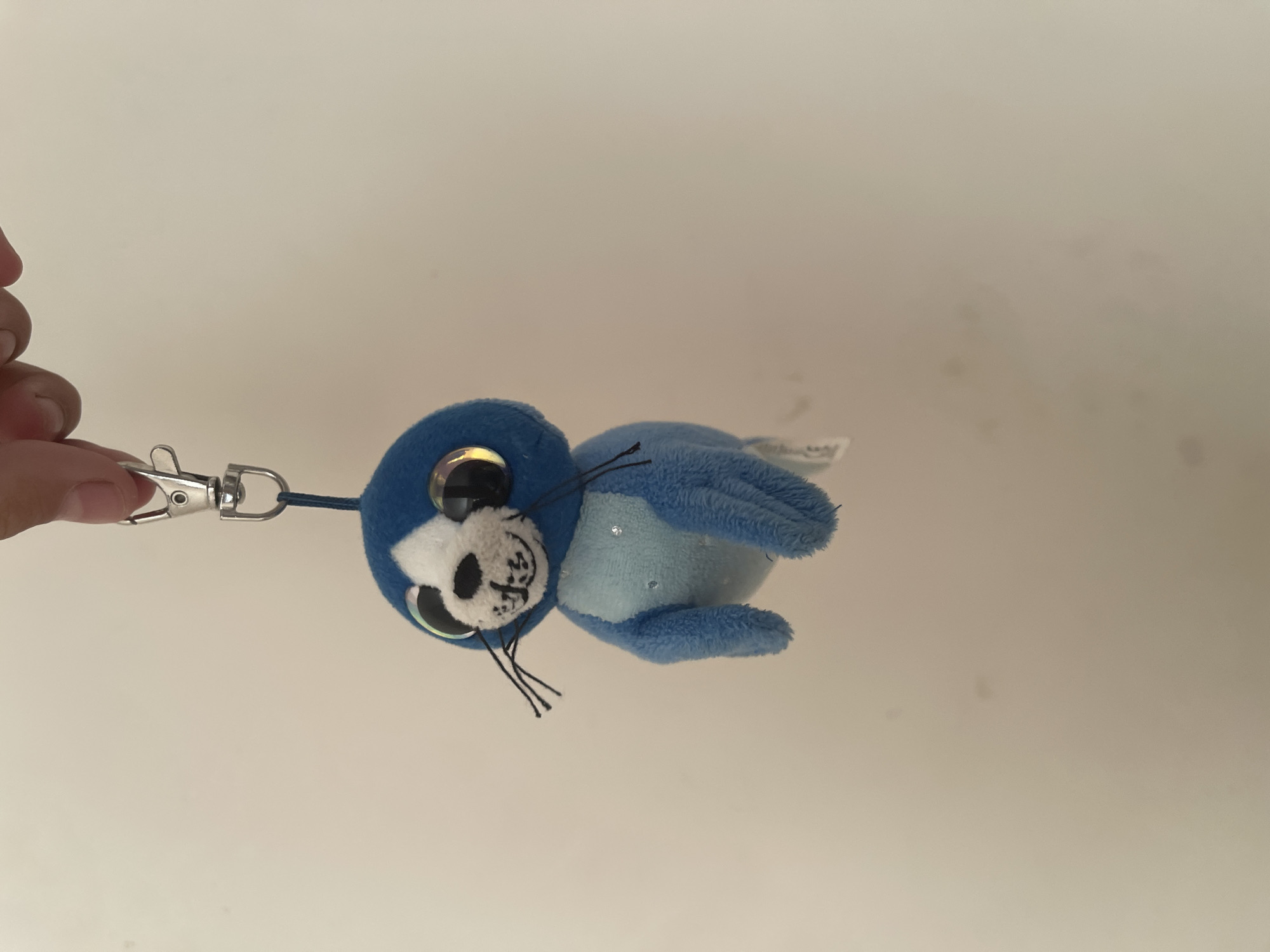 A close-up shot captures a blue plush seal keychain, dangling from a silver clasp held by a human hand. The seal, with large, iridescent eyes and black whiskers, appears to be a toy. Its body is primarily blue, with a lighter blue patch on its chest, adorned with subtle sparkles. The background is a plain, light-colored wall, suggesting an indoor setting. The location of this image is not ascertainable from the visual information provided.