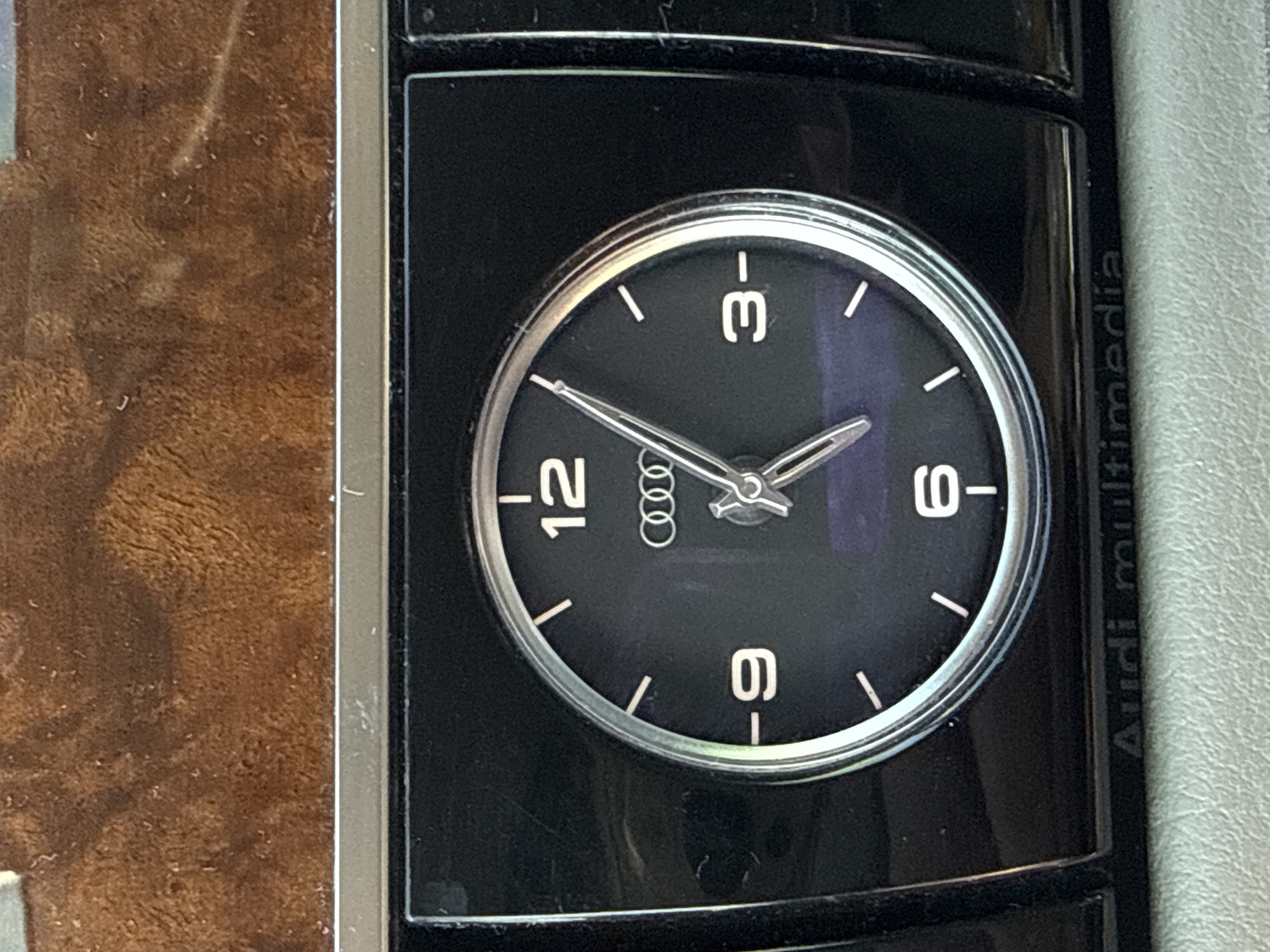 The image shows the clock of an Audi car, embedded in the dashboard. The clock face features numerals 12, 3, 6, and 9, along with the Audi logo at the center. The time displayed is approximately 1:50. The words "Audi multimedia" are visible on the right side of the clock's casing. The background shows car dashboard and the interior is made of wooden trim. There is no information available about the image location in Blagoevgrad, Bulgaria.