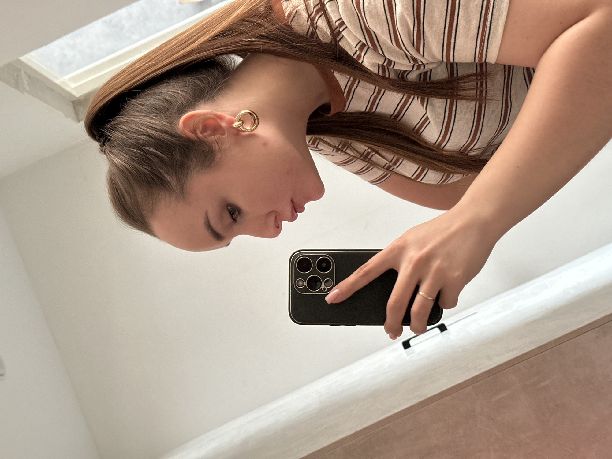 A woman takes a selfie in a bathroom. She is wearing a striped shirt and gold hoop earrings. She is holding a black smartphone with multiple cameras. The background consists of white walls and a partially visible window. There is a countertop visible at the bottom of the frame. There is no information to determine if this image was taken in the city of Chaikovsky, Russia.