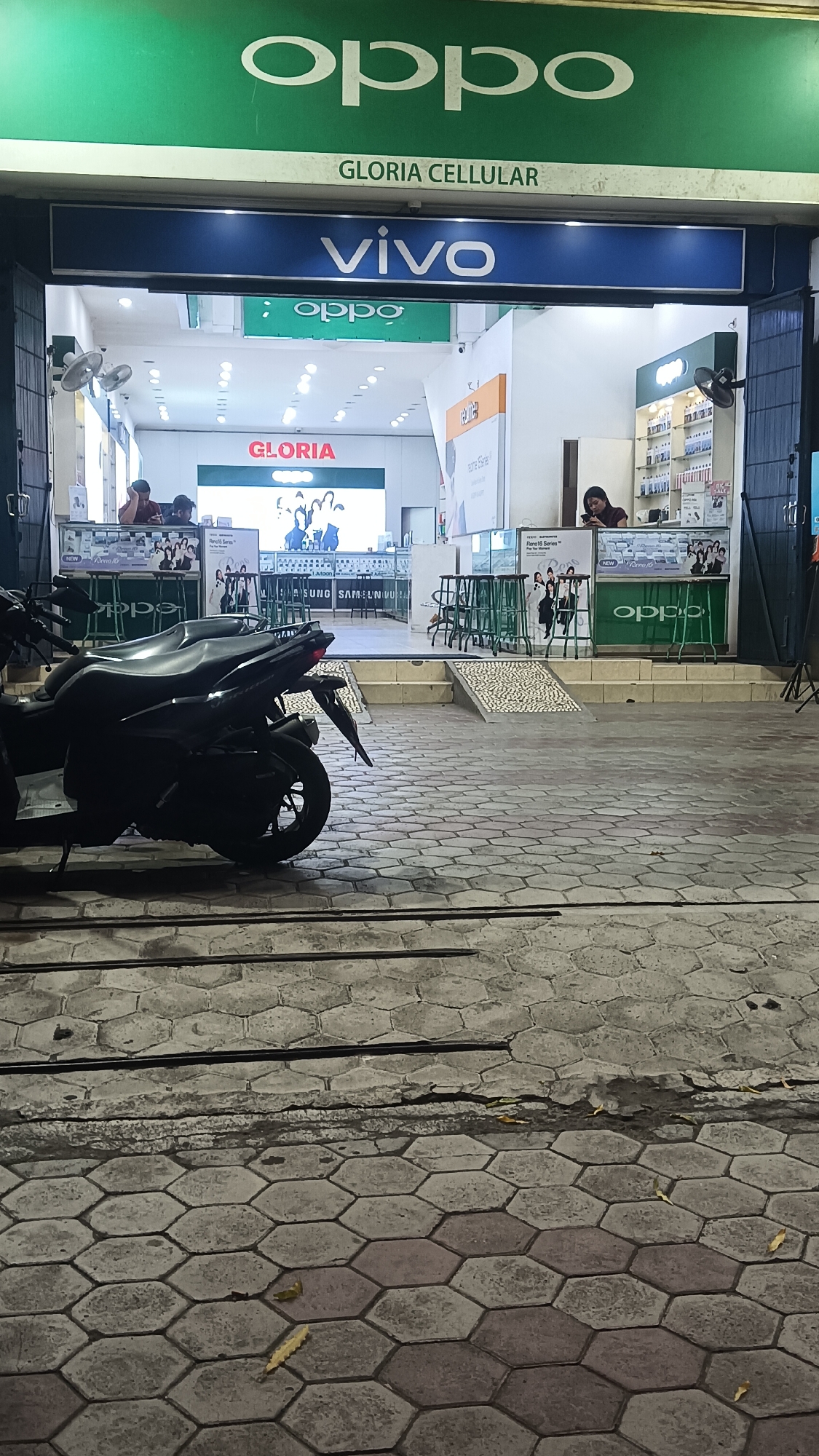 The image depicts the storefront of "GLORIA CELLULAR" in Sragen, Indonesia, at night. The store features prominent signage for OPPO and Vivo, indicating it sells mobile phones and related accessories. Inside the store, displays of phones and electronics are visible, with a few employees attending to customers or working behind counters. Outside the store, two motorcycles are parked on a paved walkway with a hexagonal pattern. The overall atmosphere is that of a typical retail establishment in an urban or semi-urban setting during evening hours.