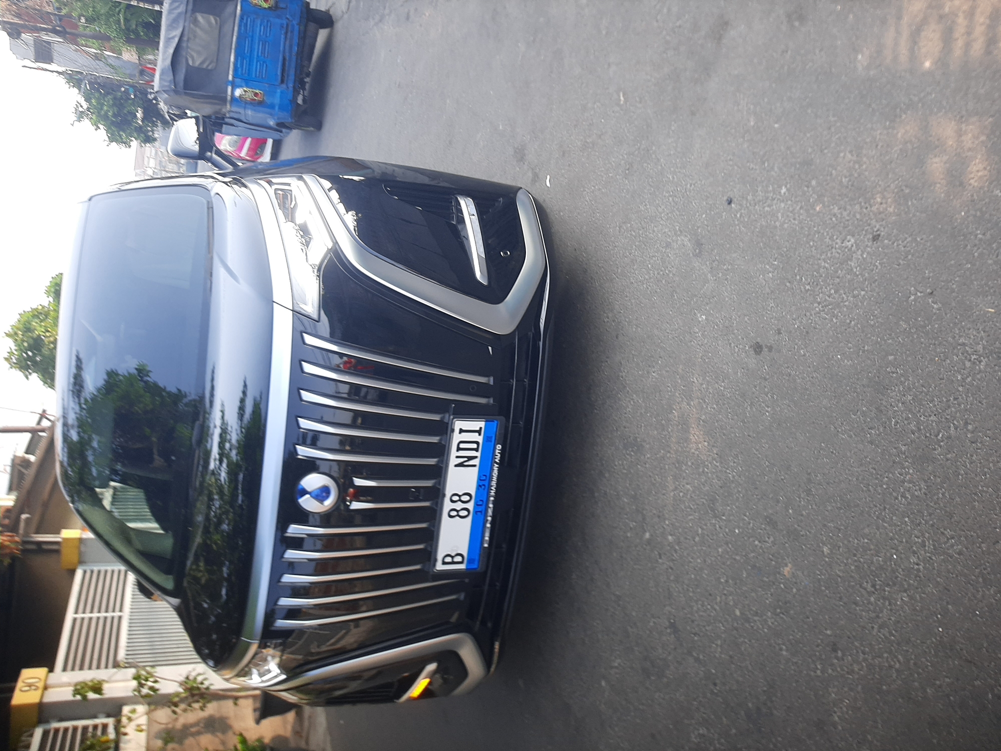 A black minivan is parked on a paved street in a residential area. The minivan has a prominent chrome grille and a blue license plate that reads "B 88 NDI". To the right of the minivan, a blue three-wheeled auto-rickshaw is visible. In the background, a house with a white fence and the number "90" is seen. Trees and other buildings line the street. The scene appears to be in a city, likely during daylight hours.