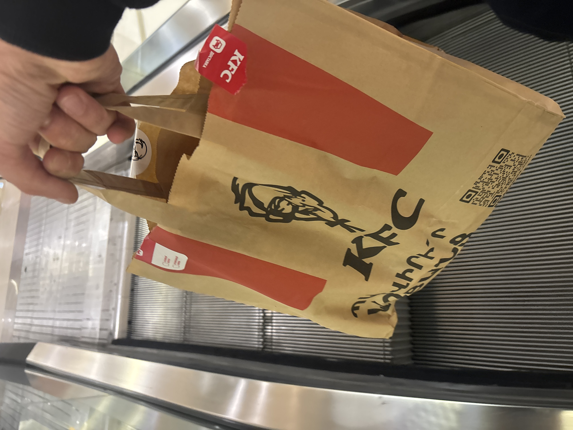 A partially visible person holds a brown paper KFC bag on an escalator. The bag has red stripes, a black KFC logo, and a red rectangular sticker with the word "KFC" printed on it. There is also a QR code printed on the bag. The escalator's metallic, grooved surface is visible beneath the bag. The photo appears to be taken at the Dalma Garden Mall in Yerevan, Armenia, based on the bag's Armenian text.