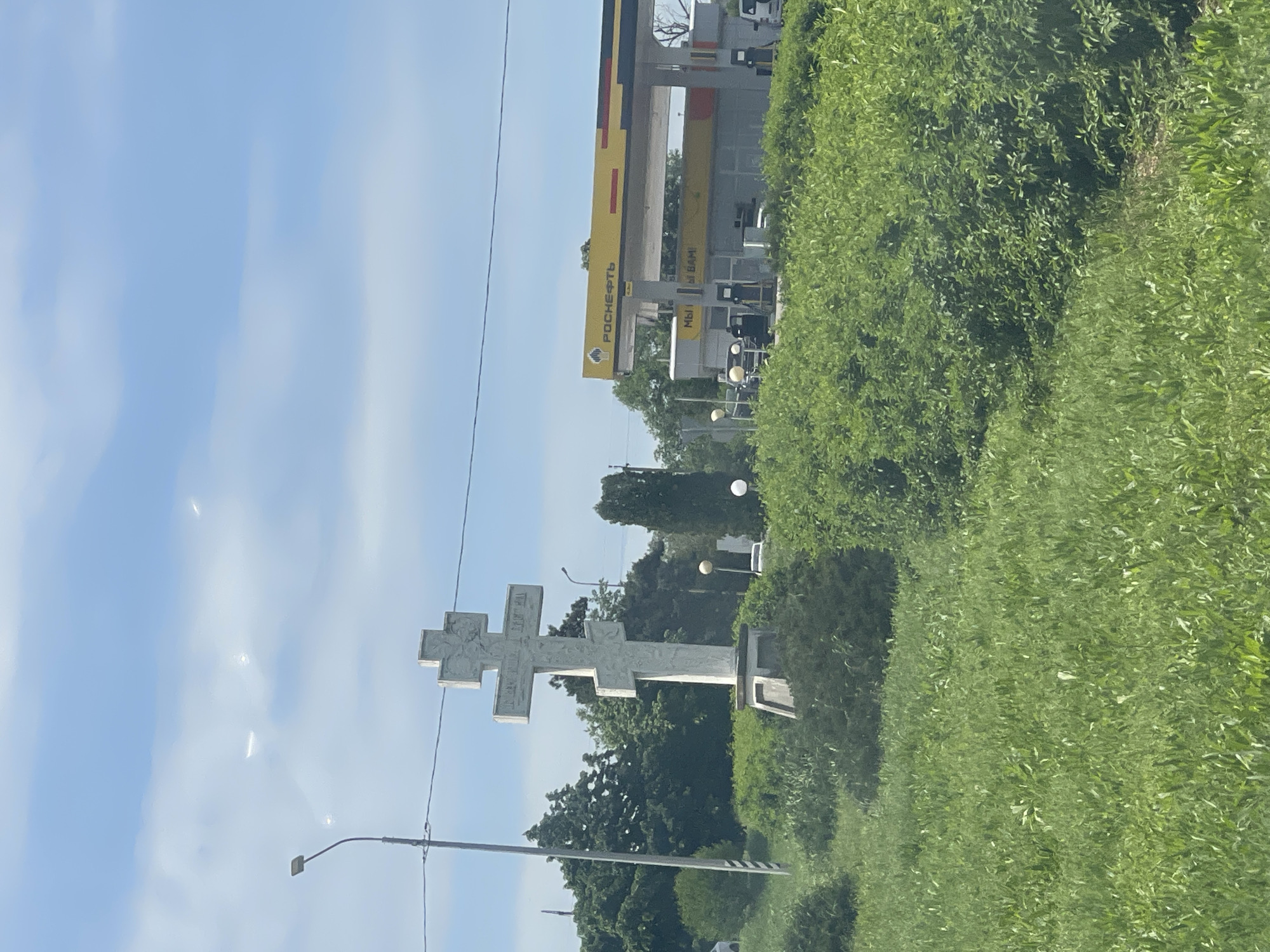 A roadside scene depicts a large white Orthodox cross erected as a monument in Donskoe, Russia, set against a backdrop of trees and a clear, blue sky. Overhead power lines and a street lamp are visible near the cross. In the distance, a "Rosneft" gas station stands at the roadside, adjacent to dense greenery.