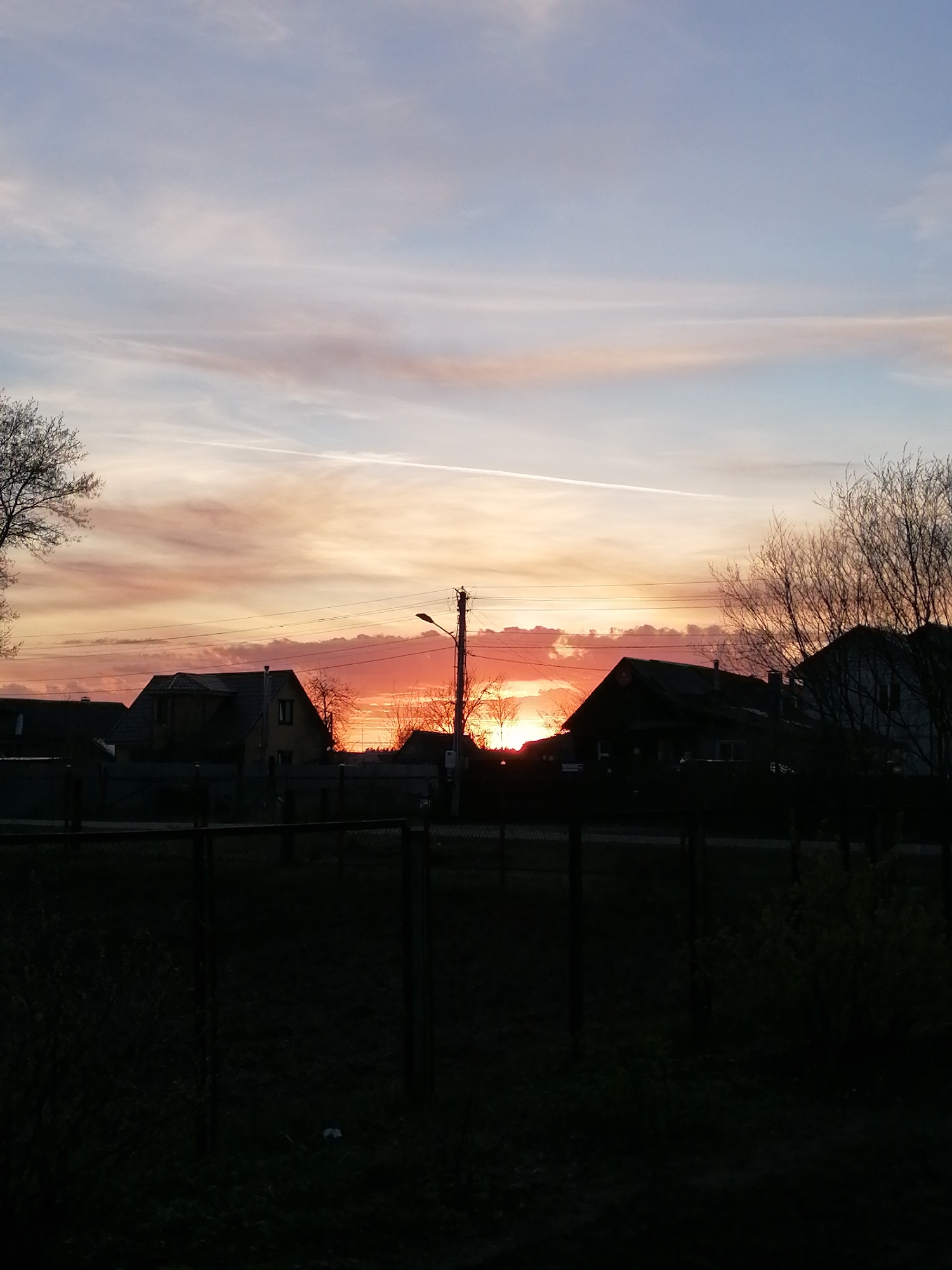 The image depicts a sunset over a residential area. Silhouetted houses are visible against the bright orange and red hues of the setting sun. Overhead, the sky transitions from orange and red near the horizon to blue and pale pink higher up, with a faint contrail stretching across the sky. A utility pole and bare trees are also silhouetted. In the foreground, a dark fence runs across the bottom of the frame. The location is identified as a residential area within the city of Unknown, Unknown.