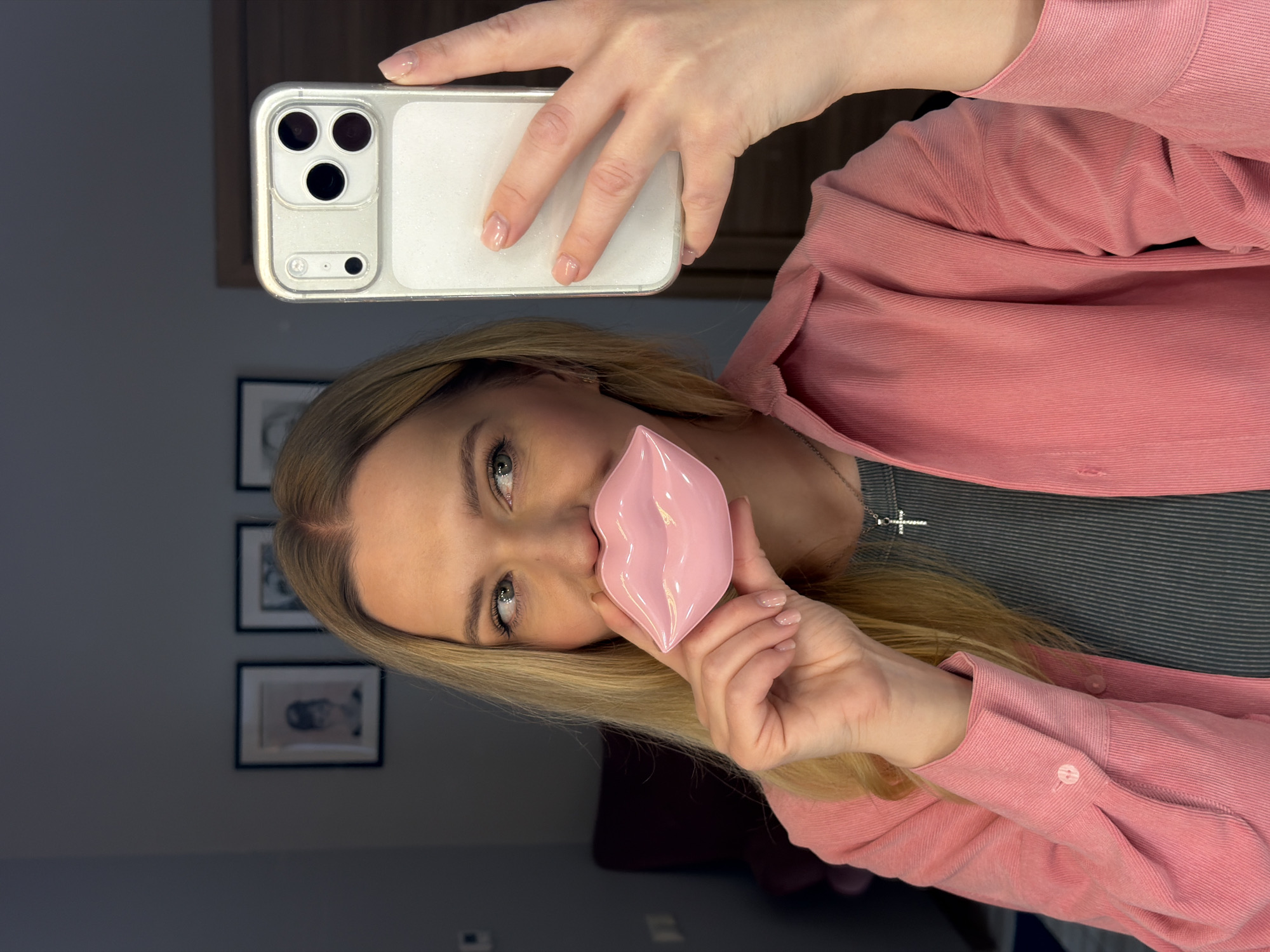 A woman is taking a selfie with an iPhone, which has a white case. She's holding a pink, lip-shaped object up to her mouth. The woman has blonde hair and is wearing a pink collared shirt over a grey shirt, and a silver cross necklace. In the background, there are framed pictures on the wall. The location is unspecified, but the city is Riga, Latvia.
