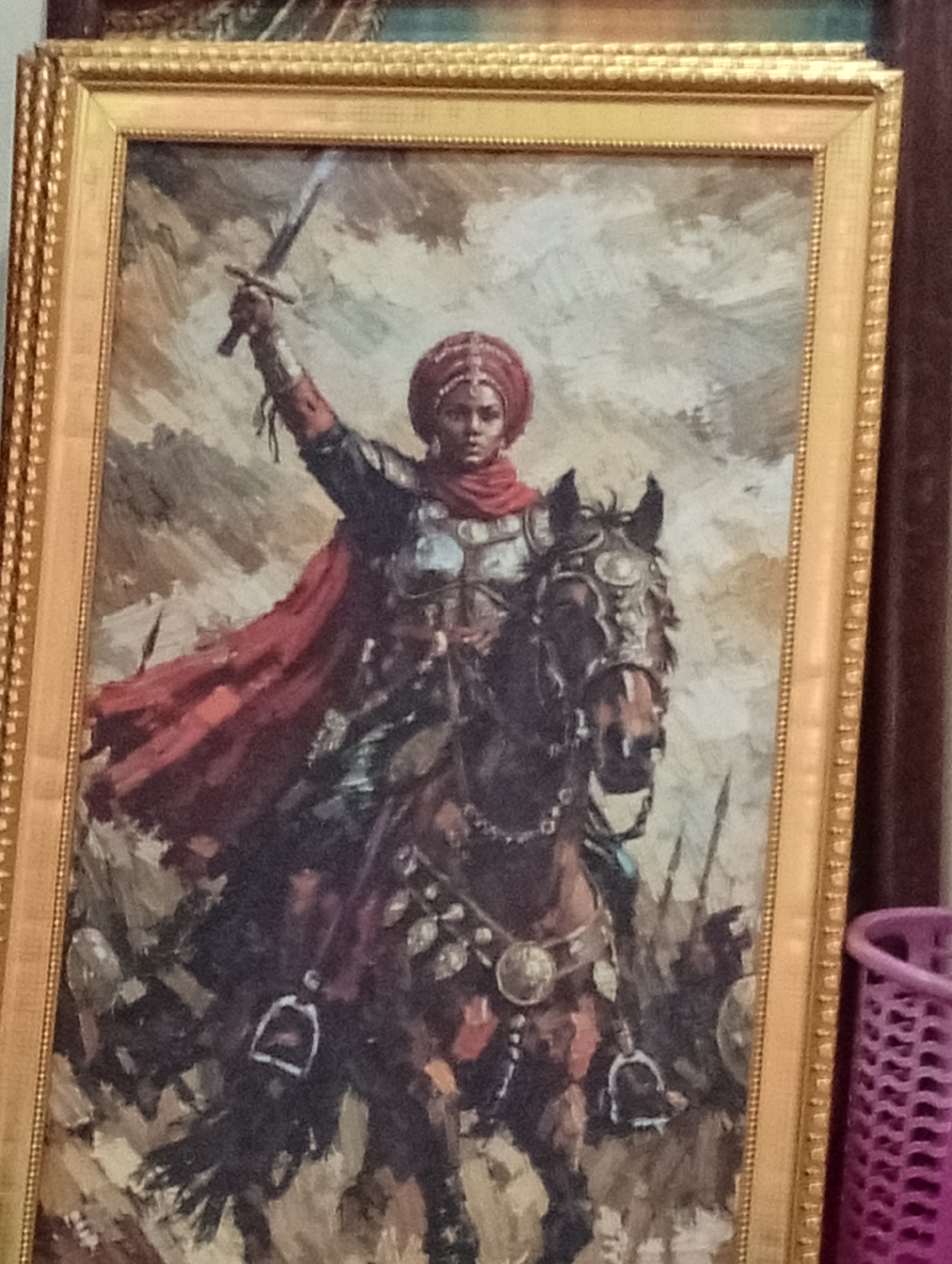 The image shows a framed painting depicting a female warrior on horseback, raising a sword. She wears a red head covering and cape, along with what appears to be armor. The horse is adorned with decorative fittings. Other figures, potentially warriors, are vaguely visible in the background. The painting is in a gold-colored frame. A purple laundry basket is partially visible to the right of the frame. The location is unknown within the city of Dadi Riba, Nigeria.