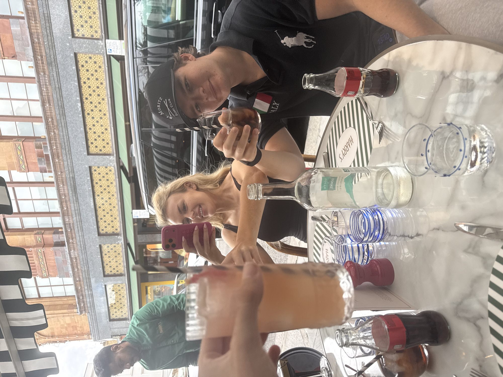 At an outdoor table at Harry's Bar in London, United Kingdom, two young adults are captured in a candid moment. A woman with blonde hair, wearing a black tank top, holds up a pink smartphone, seemingly taking a photo or video. Beside her, a young man wearing a black polo shirt and a black baseball cap with "ST. MORITZ SUSHI CLUB" on it, raises a glass of cola with ice and a straw. In the foreground, a hand holding a tall glass of pink lemonade is visible. The table is adorned with a marble-like surface and a menu with green and white stripes. Various drinks, including a bottle of Coca-Cola and a bottle of water, are arranged on the table. In the background, a waiter in a green uniform with "HARRY'S" embroidered on it is seen serving customers. The scene is set against the backdrop of a building with a grand facade and the striped awning of the establishment.