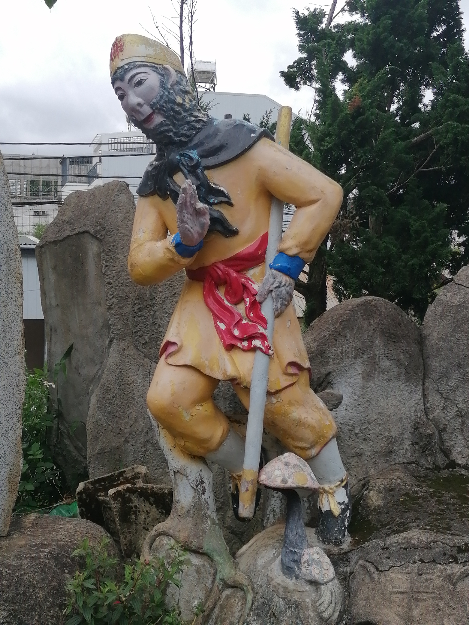 The image shows a statue of Sun Wukong, the Monkey King, in front of large rocks and greenery. The statue is brightly colored, with yellow robes, a red sash, and a golden hat. He is holding a staff and has a gesture of greeting with his open hand. In the background, there are buildings and trees. This statue is located in the garden of the Linh Phuoc Pagoda, a Buddhist temple in Da Lat, Vietnam. The temple is known for its elaborate mosaics and statues, and the Sun Wukong statue is a popular attraction.
