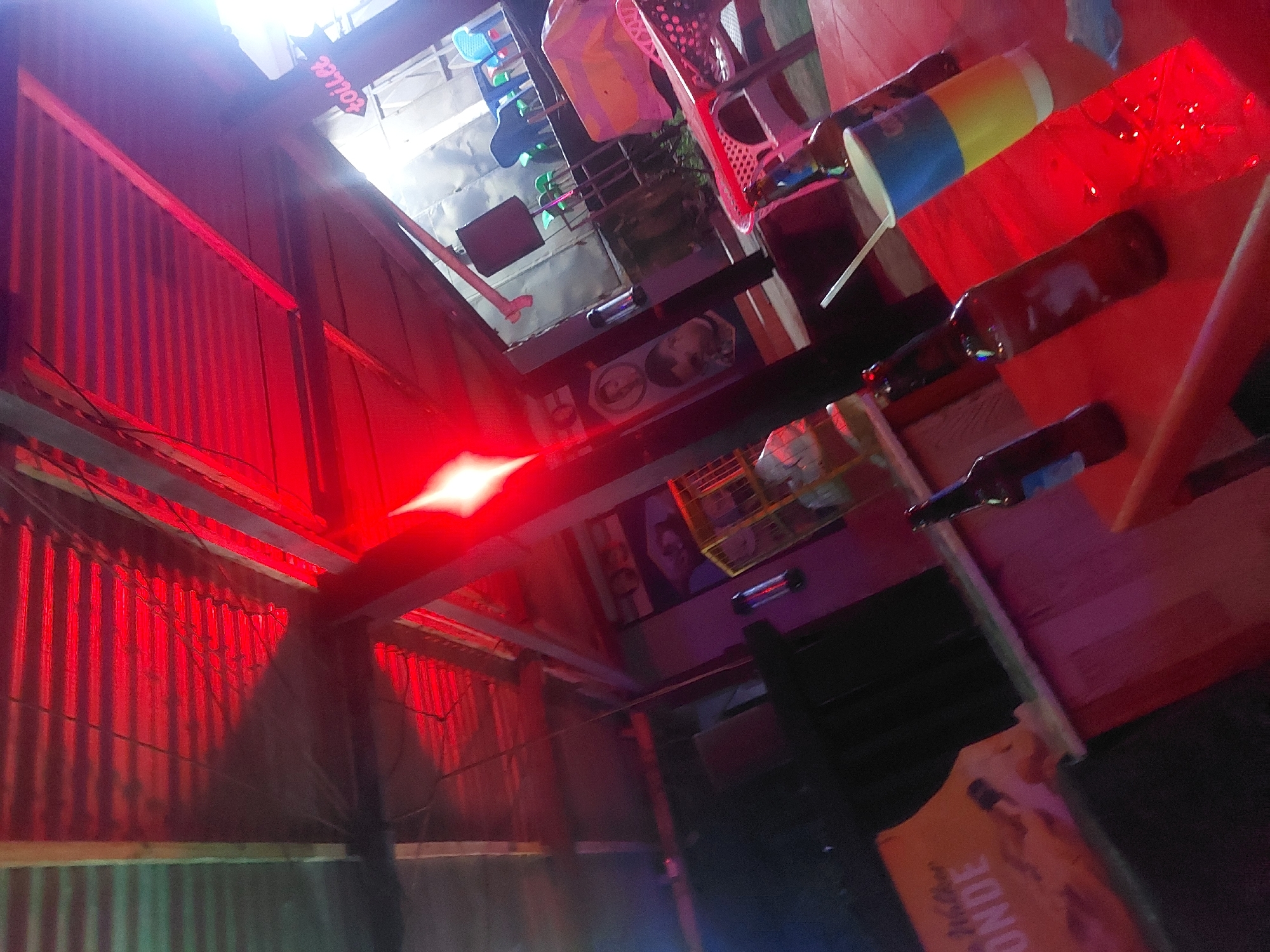 The image depicts the interior of a bar or small restaurant in Dar es-Salaam, Tanzania. Red lighting dominates the scene, illuminating corrugated metal walls. Tables are visible, with various objects placed on them including bottles, cups with straws, and a yellow sign bearing the word "BLONDE". The scene suggests an informal, possibly nighttime setting.