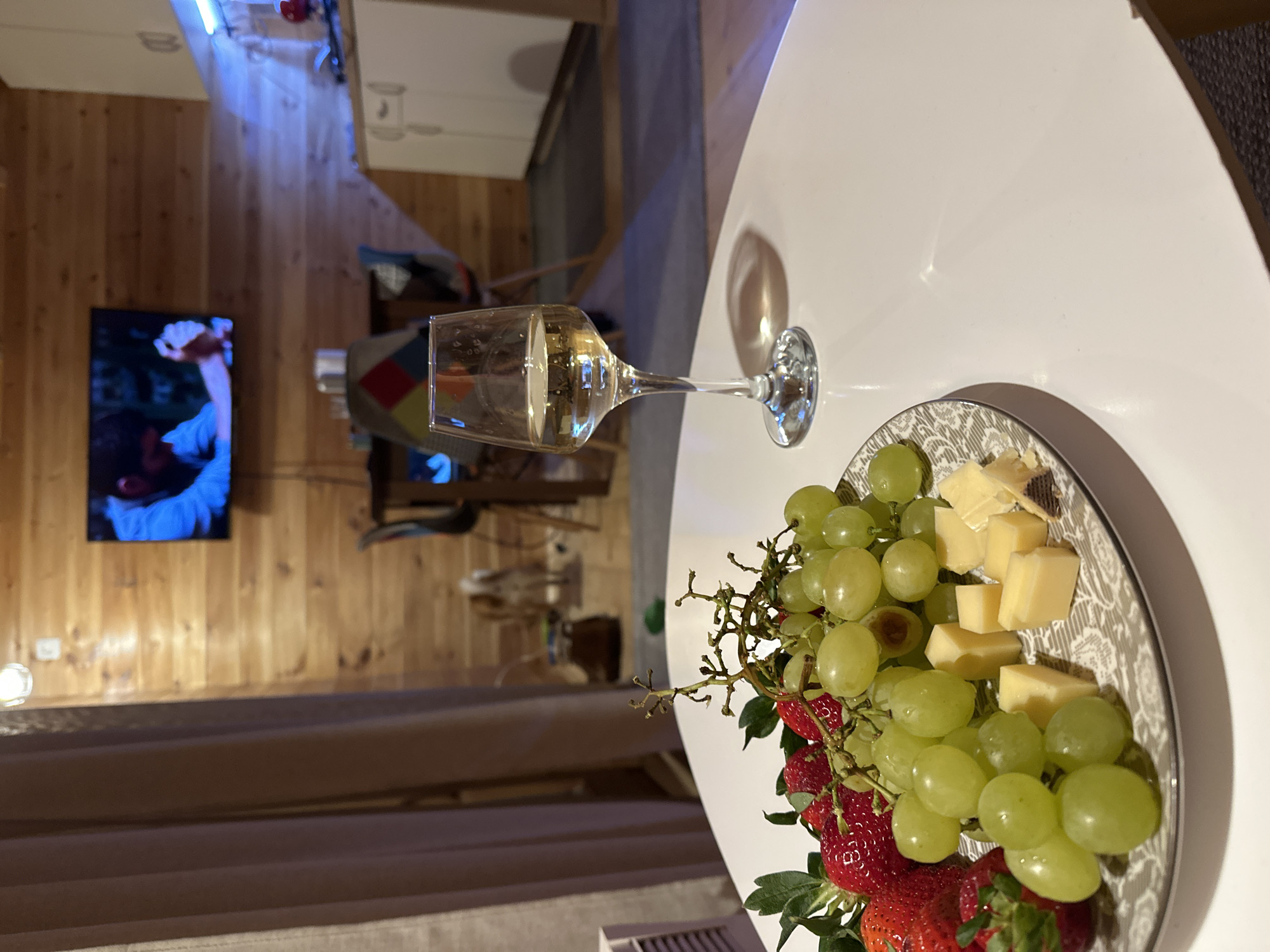 This image depicts an indoor scene in Доња Брезна, Montenegro. In the foreground, a white, rounded table displays a decorative plate laden with food and a glass. The plate contains numerous green grapes still on the vine, several red strawberries with green stems, and irregularly cut cubes of light yellow cheese. To the right of the plate, a clear wine glass, partially filled with a pale yellow liquid consistent with white wine, stands on the table.
The background reveals a room with wooden paneling on the walls. A flat-screen television is mounted on the wooden wall, displaying a scene where a male figure, wearing a light blue shirt, is visible in profile, with another male figure or part of a scene behind him. Below the television, a dining area is visible, featuring a table and at least two chairs with patterned backs. Further right, a kitchen area with white cabinets, a light-colored countertop, and illuminated blue under-cabinet lighting is present. On the floor in the mid-ground, a light brown and white dog is visible, facing away from the camera. A dark curtain is partially visible on the left side of the frame. The floor appears to be a mix of dark carpet or rug and possibly lighter flooring near the kitchen.