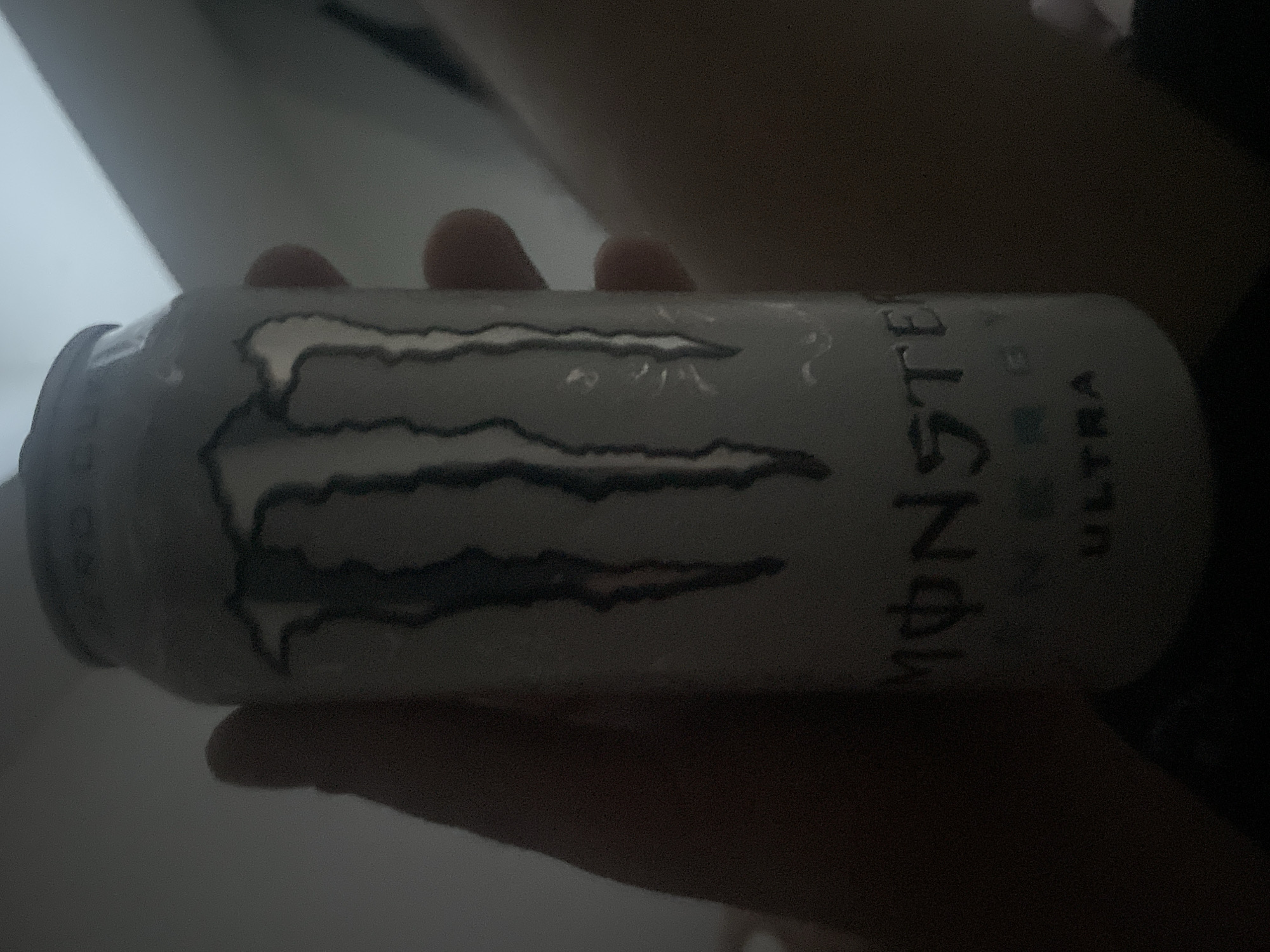A person's hand is holding a white can of Monster Energy drink. The can has the Monster Energy logo printed on it, which consists of three claw marks. Below the logo, the word "MONSTER" is visible, followed by "ENERGY" and "ULTRA". The background is dark and blurry, with some hints of light in the upper right corner, suggesting an indoor environment. The image quality is poor due to low light conditions. There is no information to suggest the location is in Paczkowo, Poland.