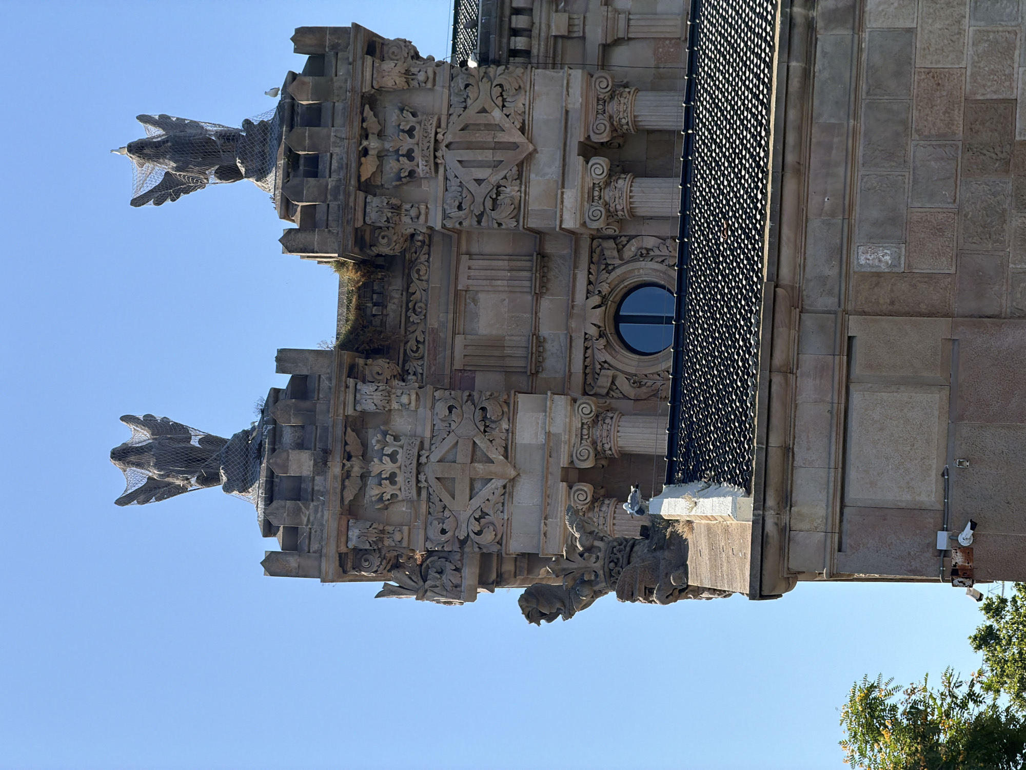 The image shows a close-up, high-angle shot of the exterior of a building with intricate stone carvings. The building appears to be historical, possibly a cathedral or a grand public building. Two gargoyle-like figures, covered in netting, protrude from the facade. One is on the left side of the image, appearing to be a dragon or a similar mythical creature with outstretched wings. The other is on the right, more difficult to discern but also appears to be a grotesque carving.
The stonework is highly detailed, with ornate patterns, architectural elements like columns with Corinthian capitals, and decorative friezes. A circular window with a blue pane is visible on the right side of the building, set within the elaborate carvings. The building's facade is constructed of stone blocks. A black metal mesh runs along the upper right portion of the building, possibly a protective covering or part of the roof structure.
The sky is a clear, bright blue, suggesting a daytime scene with fair weather. The image is rotated 90 degrees clockwise, meaning the top of the building is to the right of the frame and the ground is not visible. Two security cameras are mounted on the wall on the far right side of the building. A small bird is perched on the netting of the gargoyle on the right. A lush green tree with yellow flowers is visible in the lower right corner of the image, providing a hint of the surrounding environment. The location context identifies the scene as Barcelona, Spain. There is no visible text in the image.
