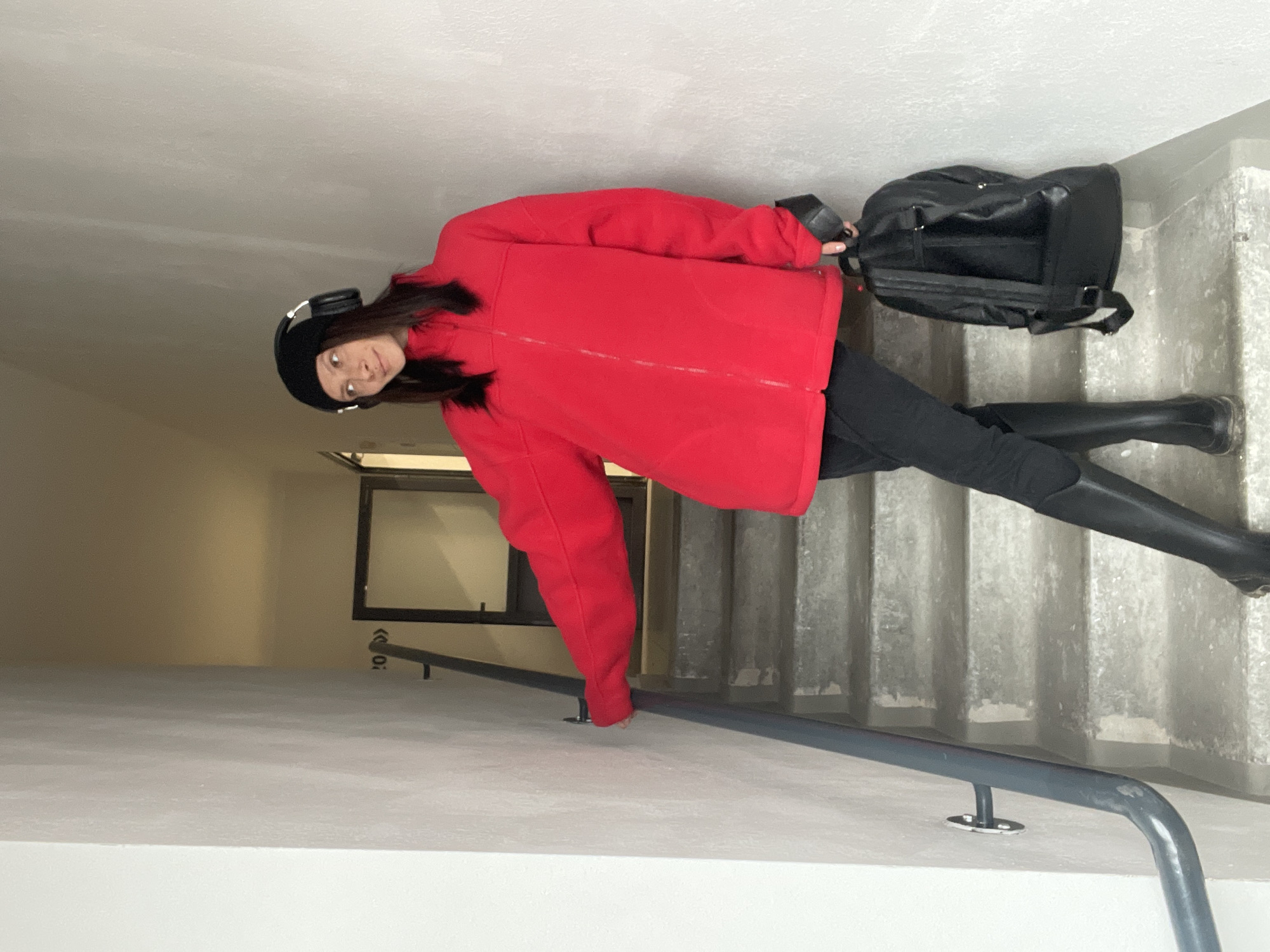 A young woman stands on a concrete staircase in Izhevsk, Russia, wearing a black beanie with headphones, a red jacket, black pants, and tall black boots. She carries a black backpack. The scene includes a window with bars, a gray metal railing, and textured concrete steps.