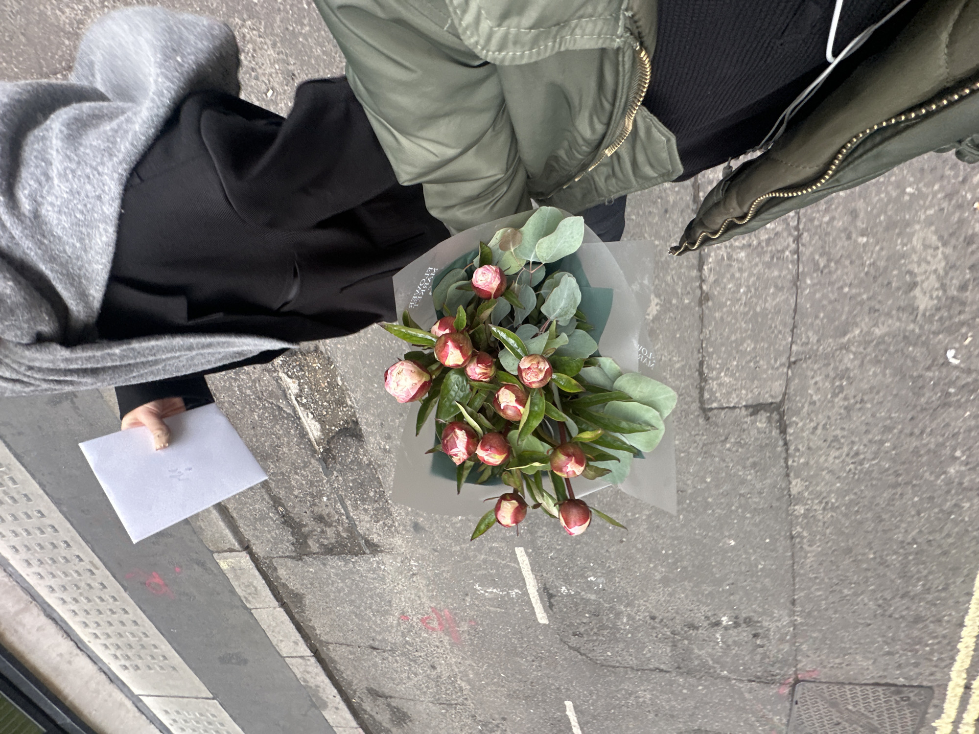 An overhead shot depicts a person on a city street, holding a bouquet of pink and white flowers wrapped in grey cellophane and a small, white envelope. The person is wearing a dark jacket, a light green bomber jacket, and a grey scarf or shawl. The bouquet is comprised of a variety of flowers with lush green leaves. The location is suggested to be a street in London, United Kingdom, inferred from the road markings and pavement. The scene is viewed from directly above, with the person and the objects they are holding in clear focus against the grey street background.