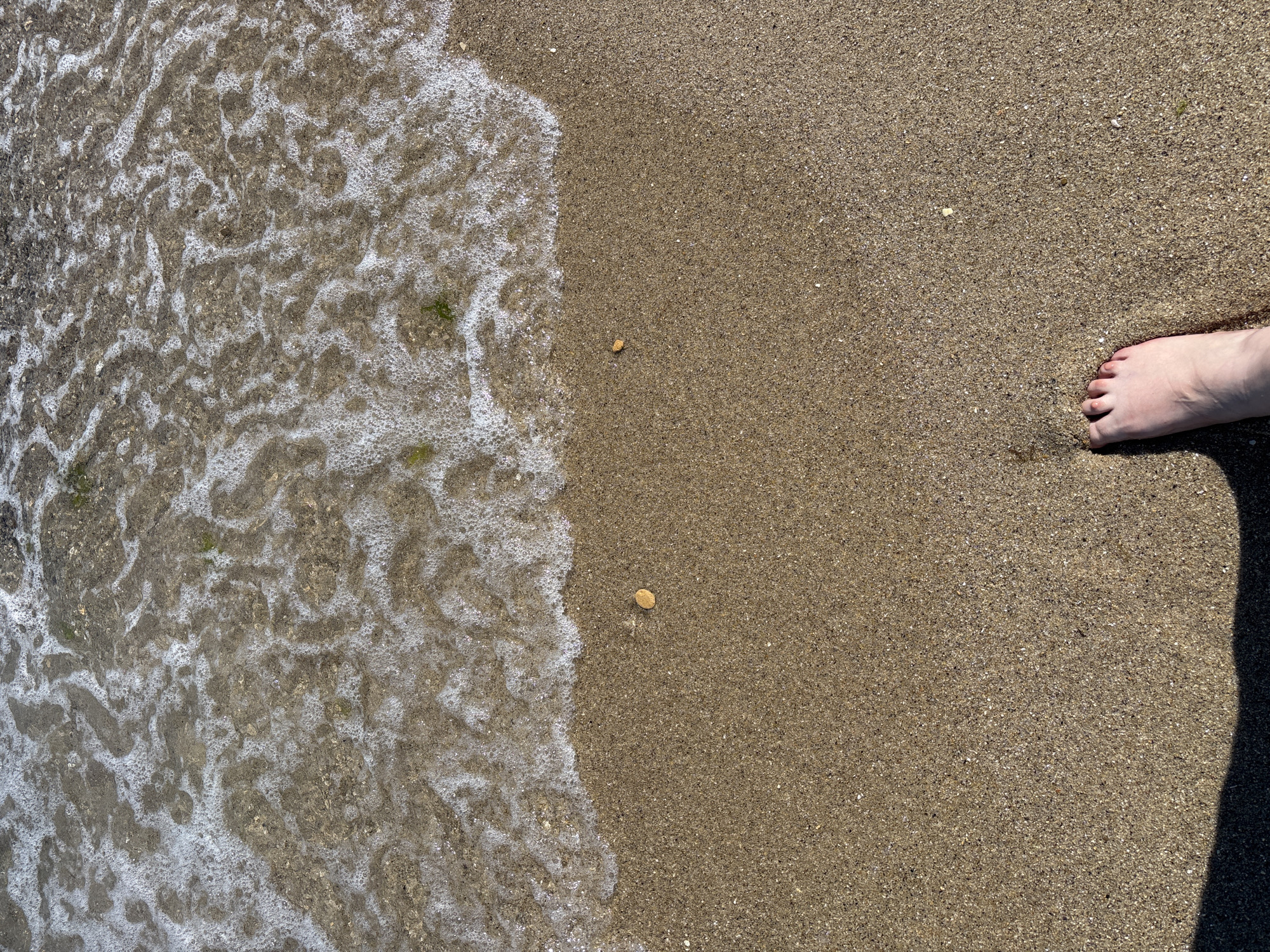 A close-up, overhead view of a sandy beach where the gentle waves of the sea are washing ashore. The water is clear and creates a frothy white foam as it recedes, revealing the wet sand underneath. Two small, light-brown pebbles are visible on the dry sand. In the lower right corner of the frame, a person's bare foot, with visible toes and skin, is positioned on the sand, casting a shadow to the left. The sand appears to be composed of small grains with a mix of light and dark particles. The image is likely taken at one of Varna, Bulgaria's many beaches, such as the Central Beach or the beach near the Sea Garden, where such sandy shorelines are common.