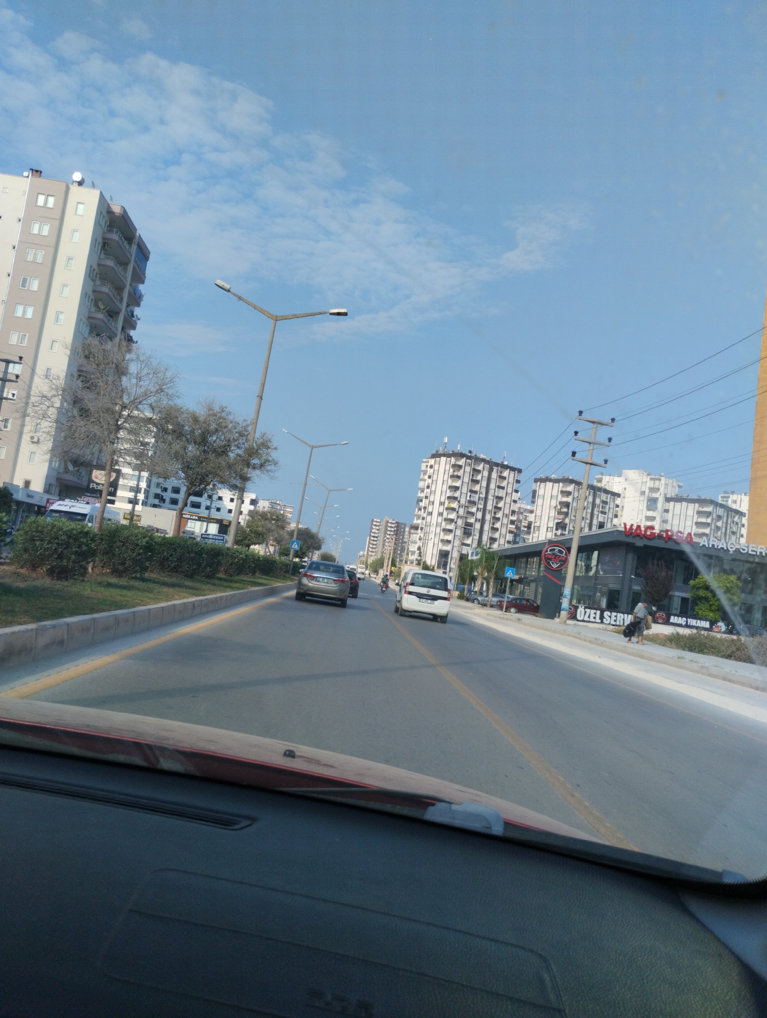 The image captures a street scene in Mezitli, Turkey, on a bright, clear day with a partly cloudy sky. The perspective is from inside a vehicle, looking forward and slightly to the right.
Dominating the left side of the frame is a tall, multi-story apartment building with a light-colored facade, numerous windows, and balconies. In front of it, bare-branched trees stand next to a row of well-maintained green bushes lining the sidewalk. Streetlights with multiple arms are visible along the road.
The road itself is paved, with a yellow line marking the lane division. Several cars are driving on the road. A silver sedan is in the foreground, followed by a white van, and further down, another car. A motorcycle is also visible in the distance.
On the right side of the road, a modern building with a dark facade and a large red and white sign that reads "VAG-PSA ARAÇ SERVİS" (meaning VAG-PSA Vehicle Service) is prominent. Below it, smaller signs read "ÖZEL SERVİS" (meaning Special Service) and "ARAÇ YIKAMA" (meaning Car Wash). A few pedestrians are on the sidewalk in front of this building. Another series of apartment buildings, similar in style to the one on the left, extends into the background on the right side of the street. The image shows a typical urban environment with residential and commercial structures, and active traffic.