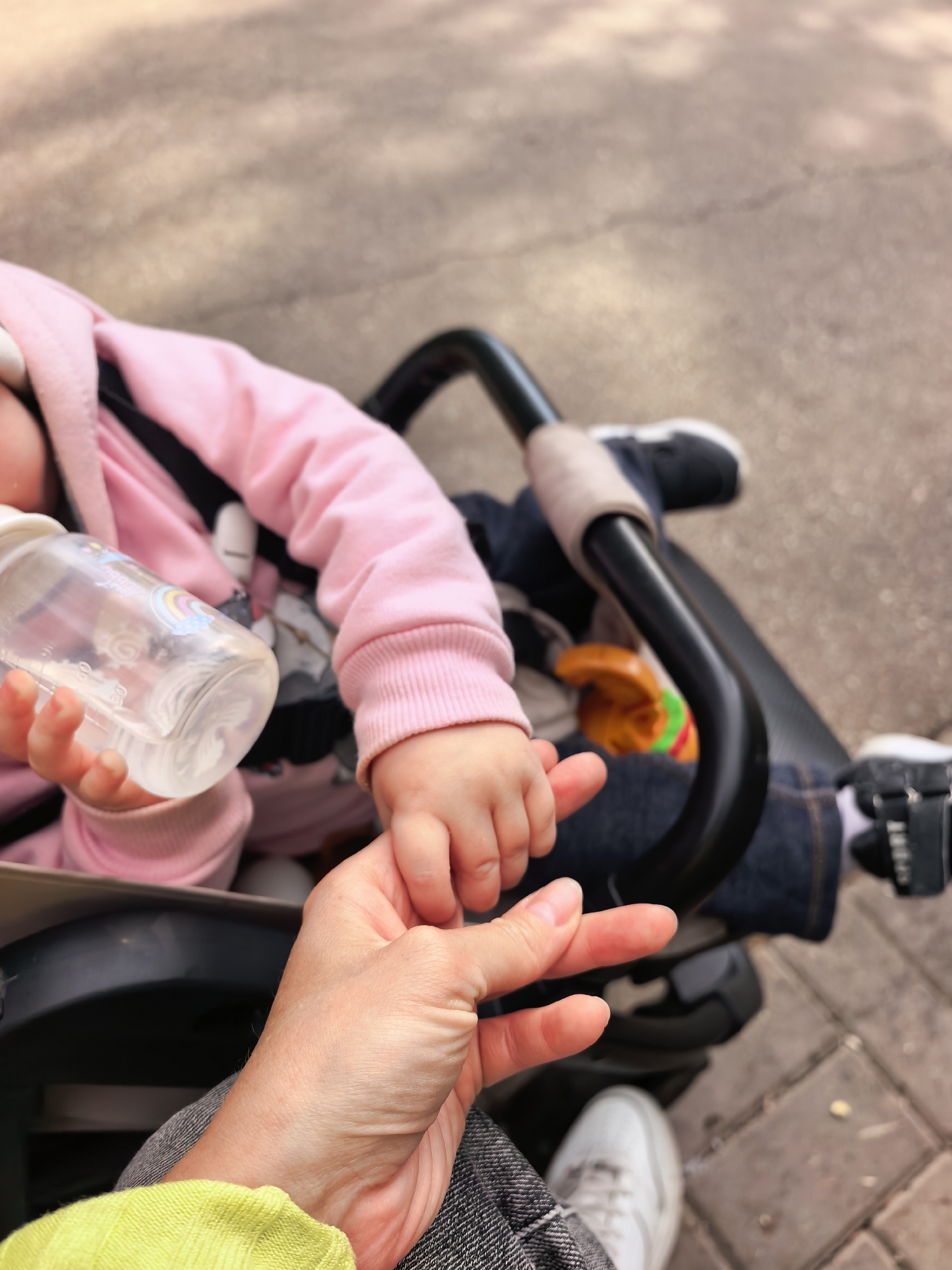 A close-up, eye-level outdoor shot captures a tender moment between an adult and a baby/toddler in Sterlitamak, Russia. The focus is on their hands and the upper body of the child, viewed from above and slightly to the side.
The baby/toddler, wearing a pink hooded top and dark jeans, is seated in a black stroller or car seat. Their small hand is gently grasped by an adult's hand, who is wearing a bright yellow sleeve and dark grey trousers. The adult's hand cradles the child's, conveying a sense of care and connection. The child holds a clear baby bottle with a white cap, filled with a transparent liquid. The bottle features volume markings (e.g., 50 ml, 100 ml, 150 ml) and a colourful rainbow and cloud design. In the background, beyond the stroller's handle, a toy with orange and green elements is visible, partially obscured.
The setting appears to be an outdoor paved area, possibly a sidewalk or pedestrian path, with light-colored concrete or cobblestones visible on the ground. The lighting suggests it is daytime, with no strong shadows indicating a specific time of day or harsh weather. The interaction between the hands is the emotional core of the image, emphasizing the bond between the two individuals.