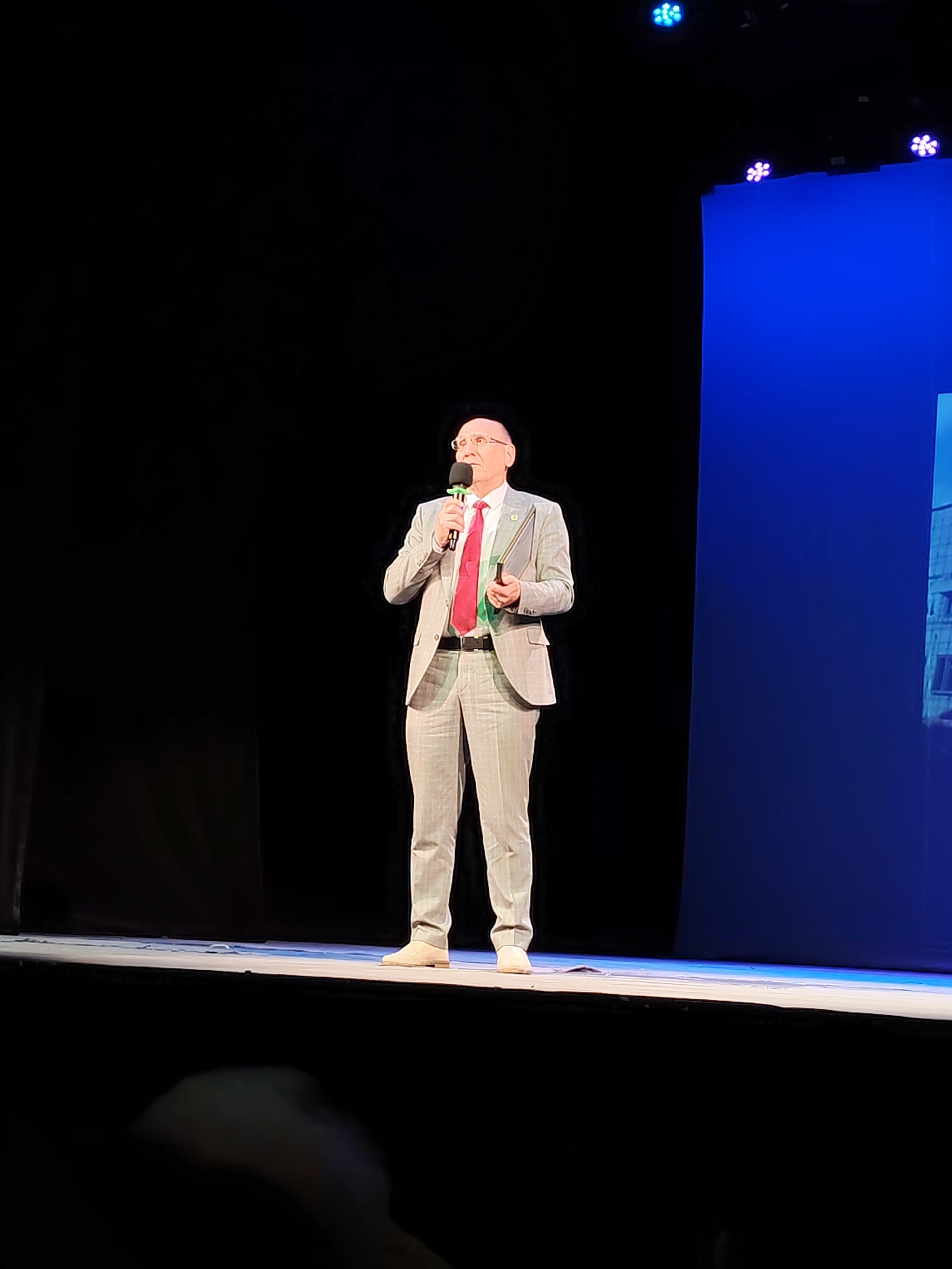 A man in a light gray suit, white shirt, and red tie stands on a stage and speaks into a microphone. He is wearing glasses and has a folder in his left hand. The stage is lit from above, with a dark background behind him and a large blue screen to his right. The image appears to be taken at the Chaikovsky State Opera and Ballet Theatre in Chaikovsky, Russia.
