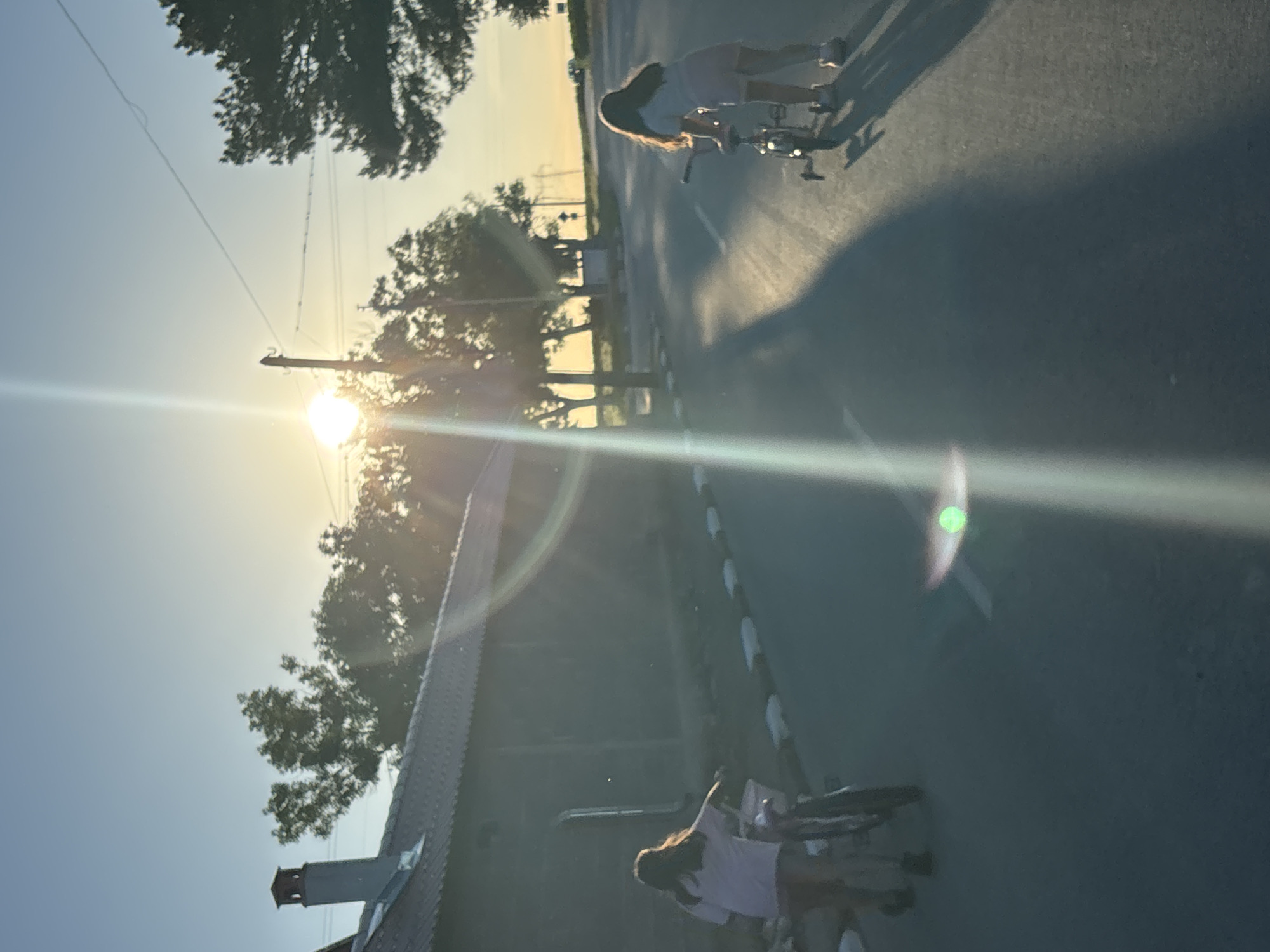 Two young girls are seen on a road in Enevo, Bulgaria, at dusk. The girl on the left, closer to a building and with her back to the camera, is pushing a bicycle. She has long, brown hair and is wearing a light-colored shirt and shorts. The girl on the right is riding a bicycle with training wheels. She also has long, brown hair and is wearing a light-colored shirt and shorts. The sun is setting, casting long shadows across the asphalt road. Trees and power lines are visible in the background, along with a building that appears to be a barn or shed. The sky is a clear blue with a faint haze from the setting sun.