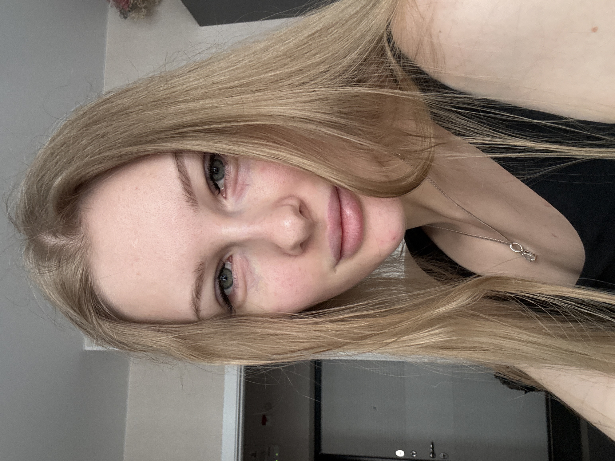 A close-up, rotated, selfie-style shot of a young woman with long, straight, blonde hair. She has fair skin, light eyes, and full lips. A delicate silver necklace is visible. The background appears to be an interior space with a gray wall and a glimpse of a black-framed doorway or piece of furniture. The image was taken in Perm, Russia.