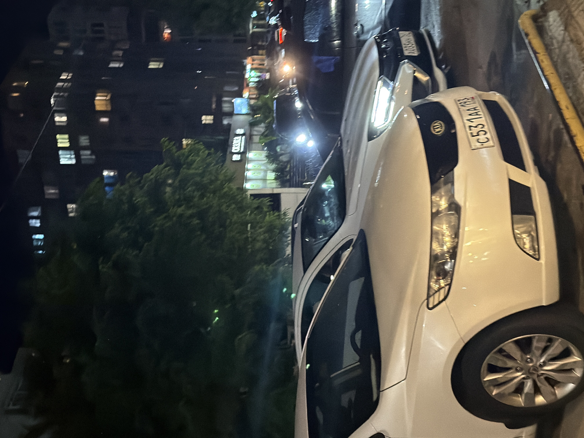 The image shows a night-time street scene in Sochi, Russia. Several cars are parked along the side of a building, which appears to be a multi-story residential or commercial building with illuminated windows. In the foreground, the front of a white car is visible, with its license plate reading "C531AA 193". Behind it, another white car is partially visible, followed by darker-colored vehicles. A tree with green foliage is prominent on the left side of the image, casting shadows. Streetlights and building lights illuminate the scene, creating a somewhat dimly lit environment. The street appears to be paved.
