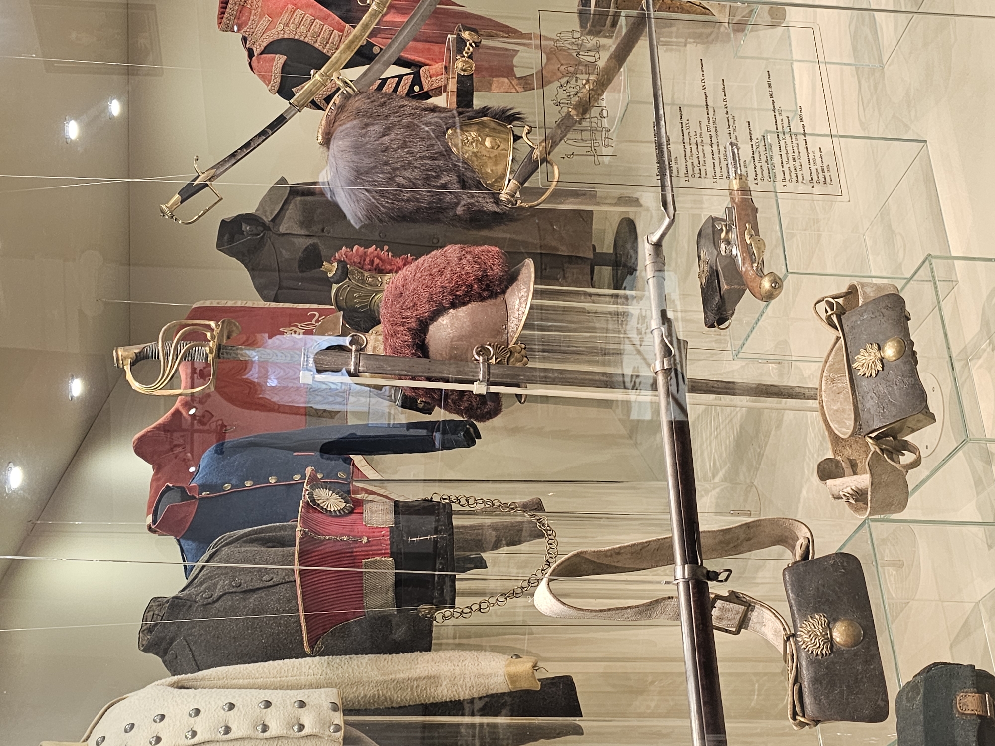 This image shows a display case filled with historical military artifacts at the Borodino Panorama Museum in Borodino, Russia. The case contains uniforms, headwear, weaponry (including swords, a rifle, and a pistol), and accompanying accessories from the Napoleonic era. The items are neatly arranged and displayed behind glass, with informational labels placed near some of the artifacts. The lighting in the museum is visible as reflections on the glass surfaces of the display case.