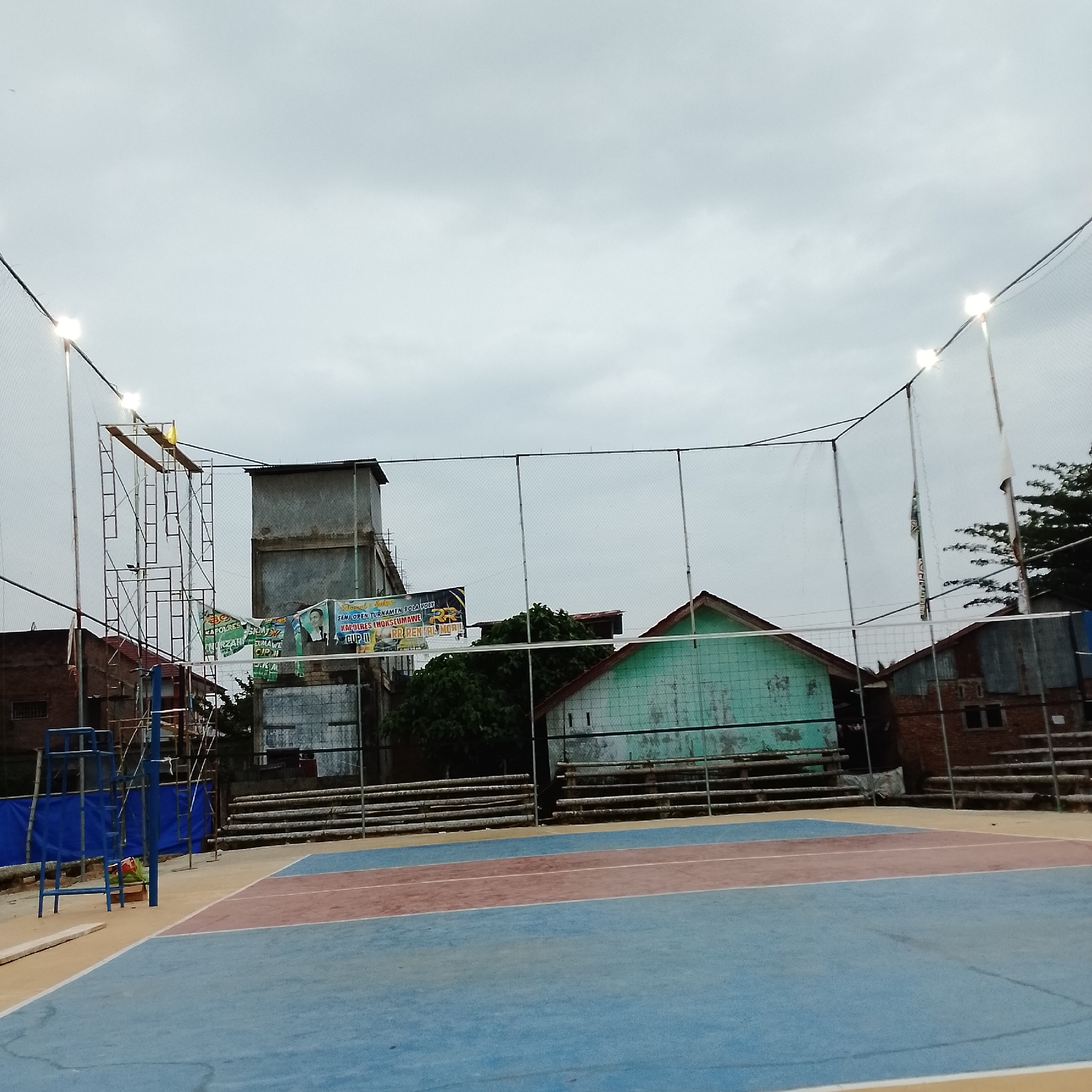 An outdoor volleyball court is depicted under a cloudy sky. The court itself is surfaced with blue and red sections. A net stretches across the width of the court, supported by poles. In the background, there are buildings, including a tall, grey structure that appears to be a water tower or utility building, and several smaller structures with various rooflines. Scaffolding is visible near the grey building. Banners are hanging from the scaffolding and from poles near the buildings. Bleachers, made of horizontal wooden planks supported by metal frames, are situated behind the court. Lights are mounted on poles around the perimeter of the court. The scene appears to be in a somewhat developed but potentially informal urban or semi-urban environment. No people are visible in the image.