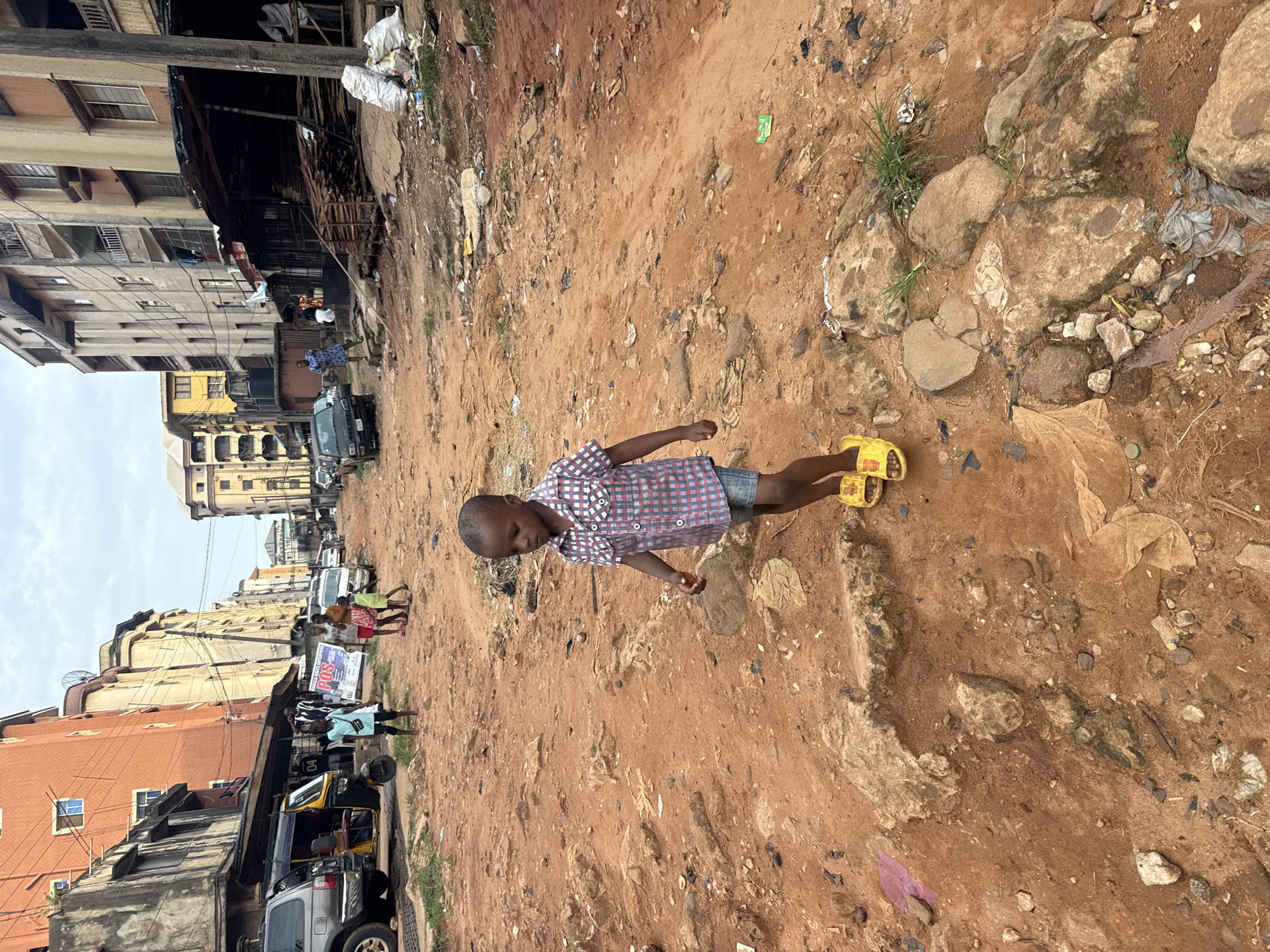 A young boy wearing a checkered shirt, denim shorts, and yellow sandals walks on a dirt road. Buildings line the street, and vehicles are parked. The scene is in Onitsha, Nigeria. The road is littered with debris, and rocks are scattered nearby. In the background, people can be seen walking and standing.
