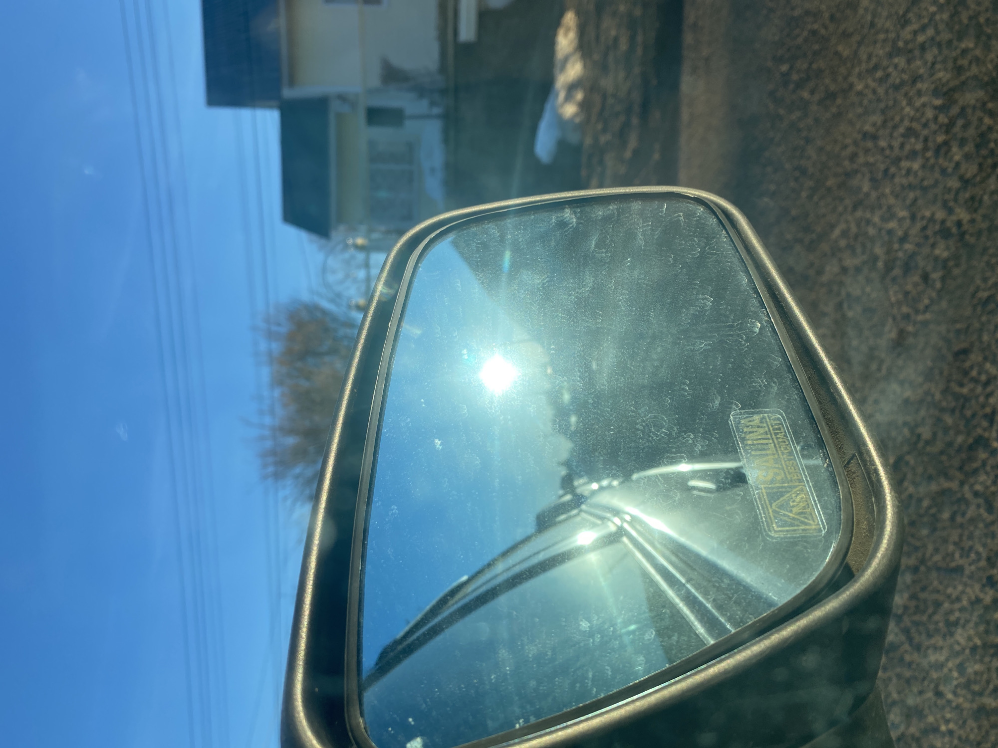 The image is a close-up of a side mirror of a vehicle, with a reflection of the surroundings. The sun is reflected in the mirror, creating a bright glare. In the background, reflected in the mirror and visible outside, is a house, bare trees, power lines, and a patch of possibly snow. The surrounding area appears to be a street or road with asphalt or a similar dark surface. There is an impression that this photograph was taken in Прикамский, Russia, since the mirror reflects the urban elements.