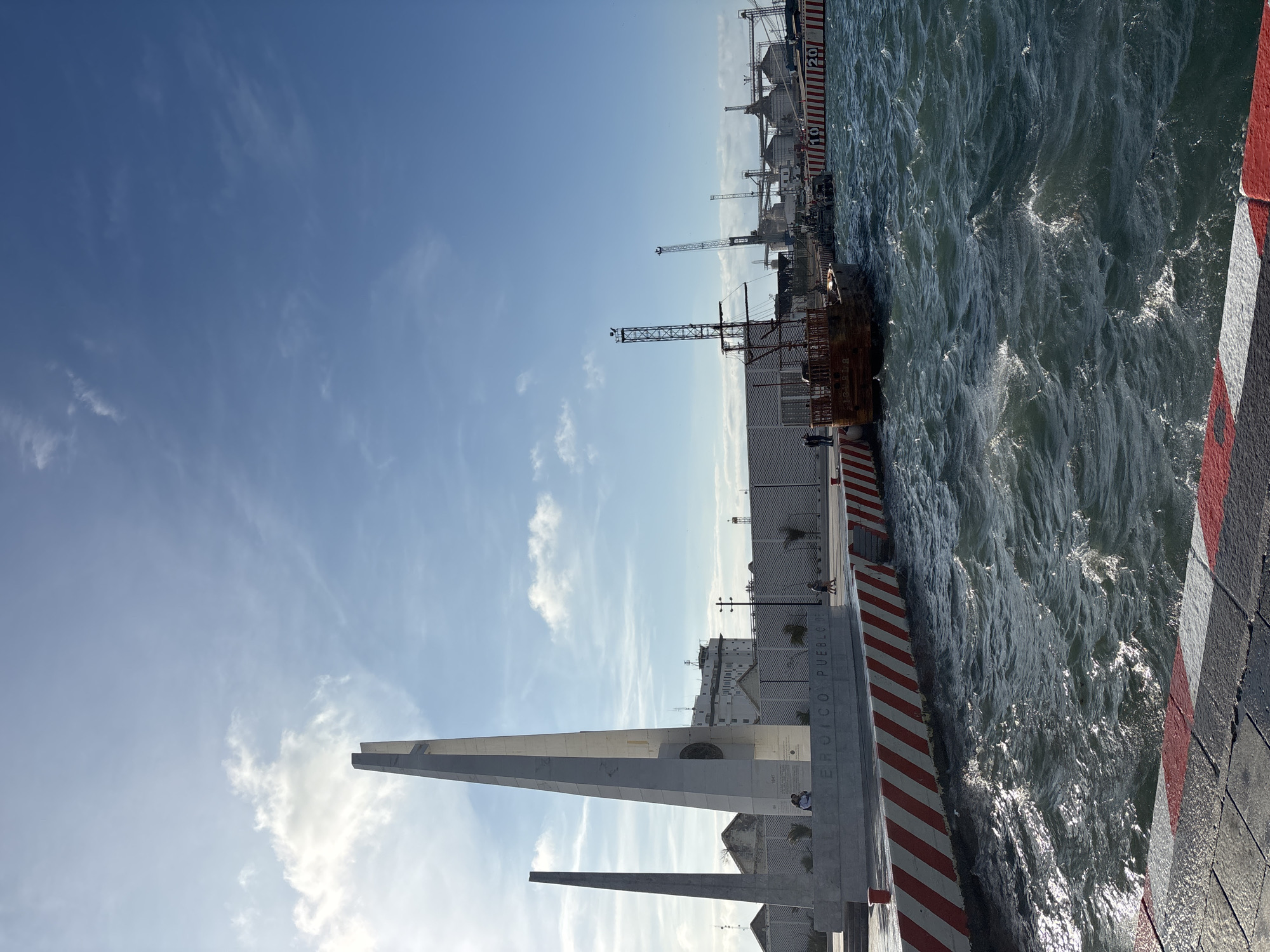 The image shows a view of the port in Veracruz, Mexico. The sky is light blue with some wispy clouds. On the left side of the frame is a white monument with a tall, triangular structure. To the right of the monument is the port, with large cranes and industrial buildings. A rusted ship is docked at the port. The water is choppy and dark blue. A red and white striped barrier runs along the edge of the port.
