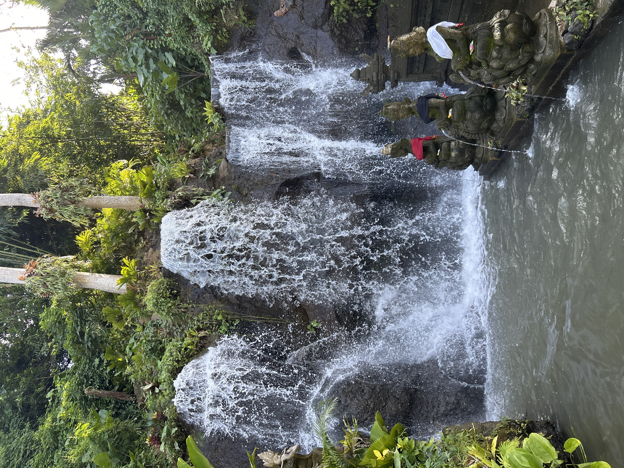 The image depicts a waterfall in Sukawati, Indonesia. The water cascades down a rocky cliff face covered in lush green vegetation. Three stone statues adorned with white, black, and red cloths stand at the edge of a pool of water at the top of the waterfall. Water pours out from the statues into the pool and down the waterfall.