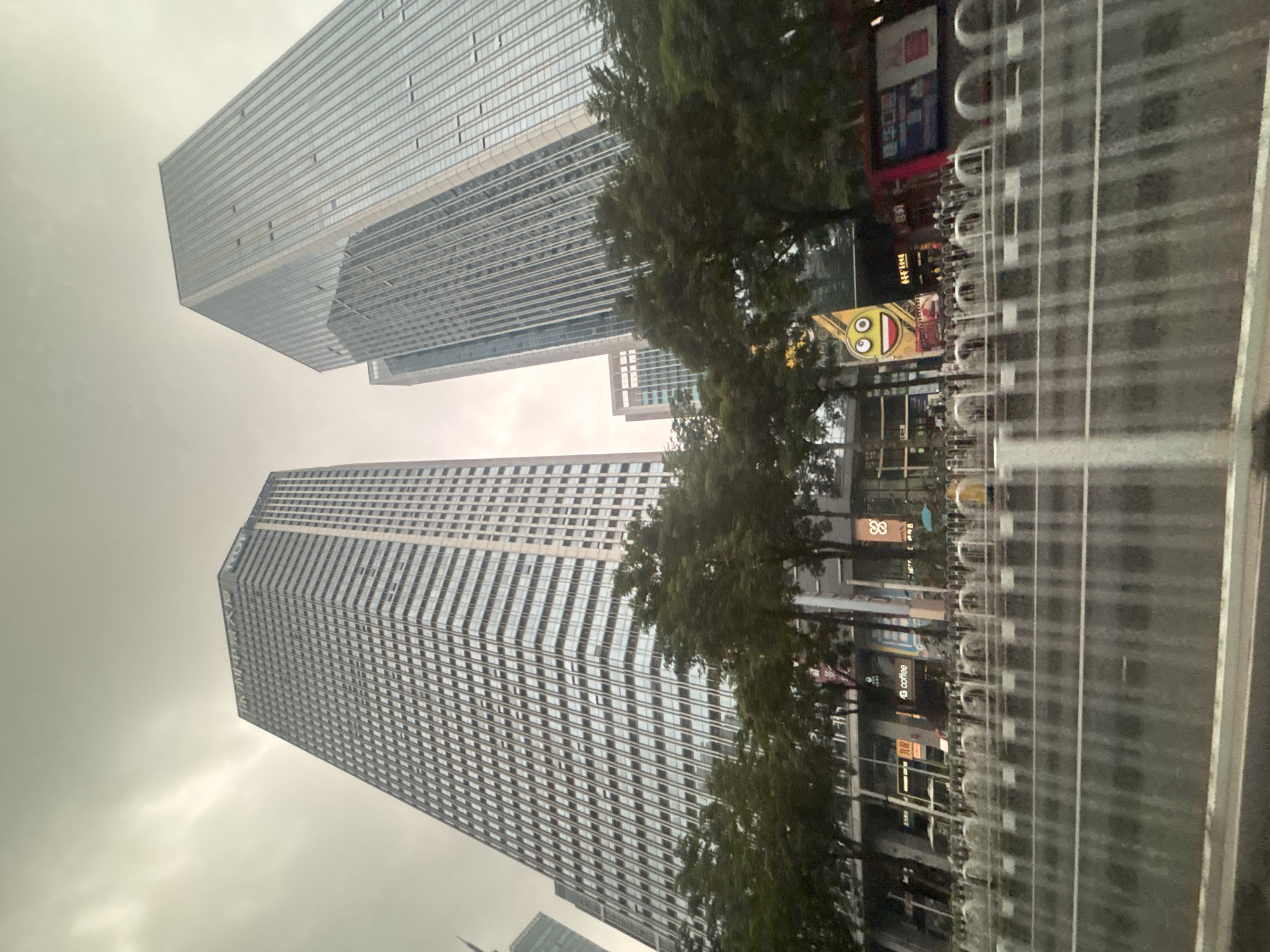 This is a low-angle, wide shot of modern skyscrapers in Tianhe District, China. The sky is overcast and grey. In the foreground, a row of white, metallic barricades is visible, with a wet road behind them. Trees line the street in front of the buildings. The buildings are tall and constructed with glass and metal facades, reflecting the cloudy sky. One building on the left has visible signage that is difficult to read clearly. To the right, another taller building with a predominantly glass exterior dominates the frame. In the mid-ground, there are storefronts with various signs and advertisements, including one with a cartoon-like emoji face. The scene appears to be a bustling urban environment.