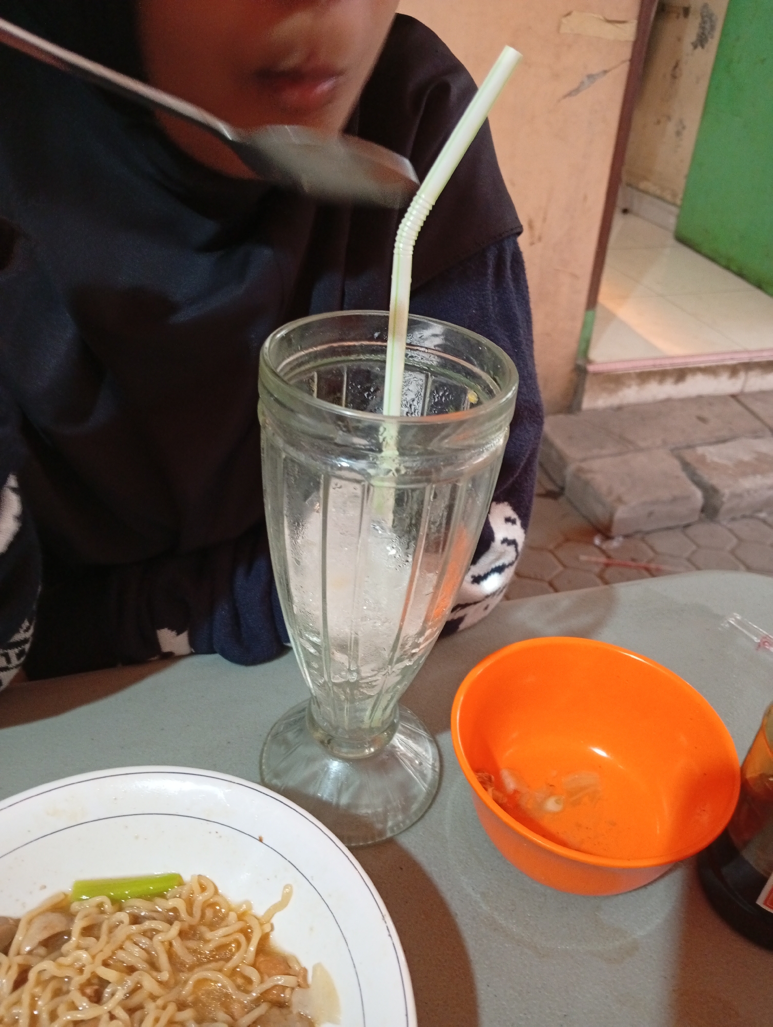 A close-up, eye-level, slightly angled shot captures a person eating noodles in Curug Wetan, Indonesia. The person, whose face is partially obscured by a spoon, is wearing a dark navy blue long-sleeved shirt and a headscarf. They are using a spoon to eat from a white bowl of noodles with green vegetables and what appear to be meatballs. To the right of the noodle bowl is a clear glass with ice and a bent white straw. Further to the right is an orange bowl with some residue inside, and part of a dark bottle is visible at the far right edge of the frame. The scene takes place outdoors, suggested by the textured concrete ground and a wall in the background, possibly a dining area in Curug Wetan.