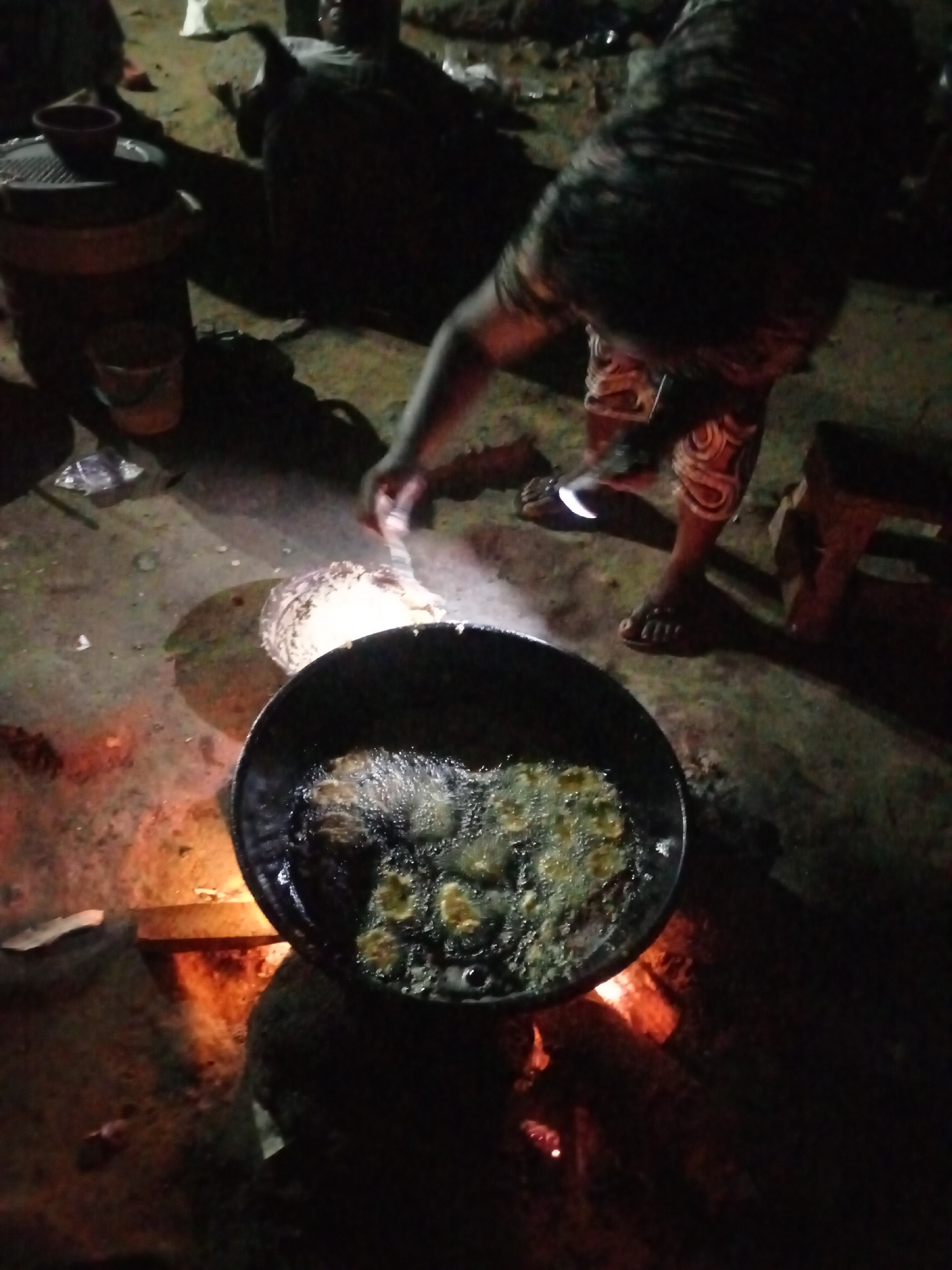A person is standing next to a large black pan filled with frying objects. The pan sits above a wood fire. To the left, there is a small pot or container. The scene is dimly lit. Based on the prompt, this is likely in Keffi, Nigeria.