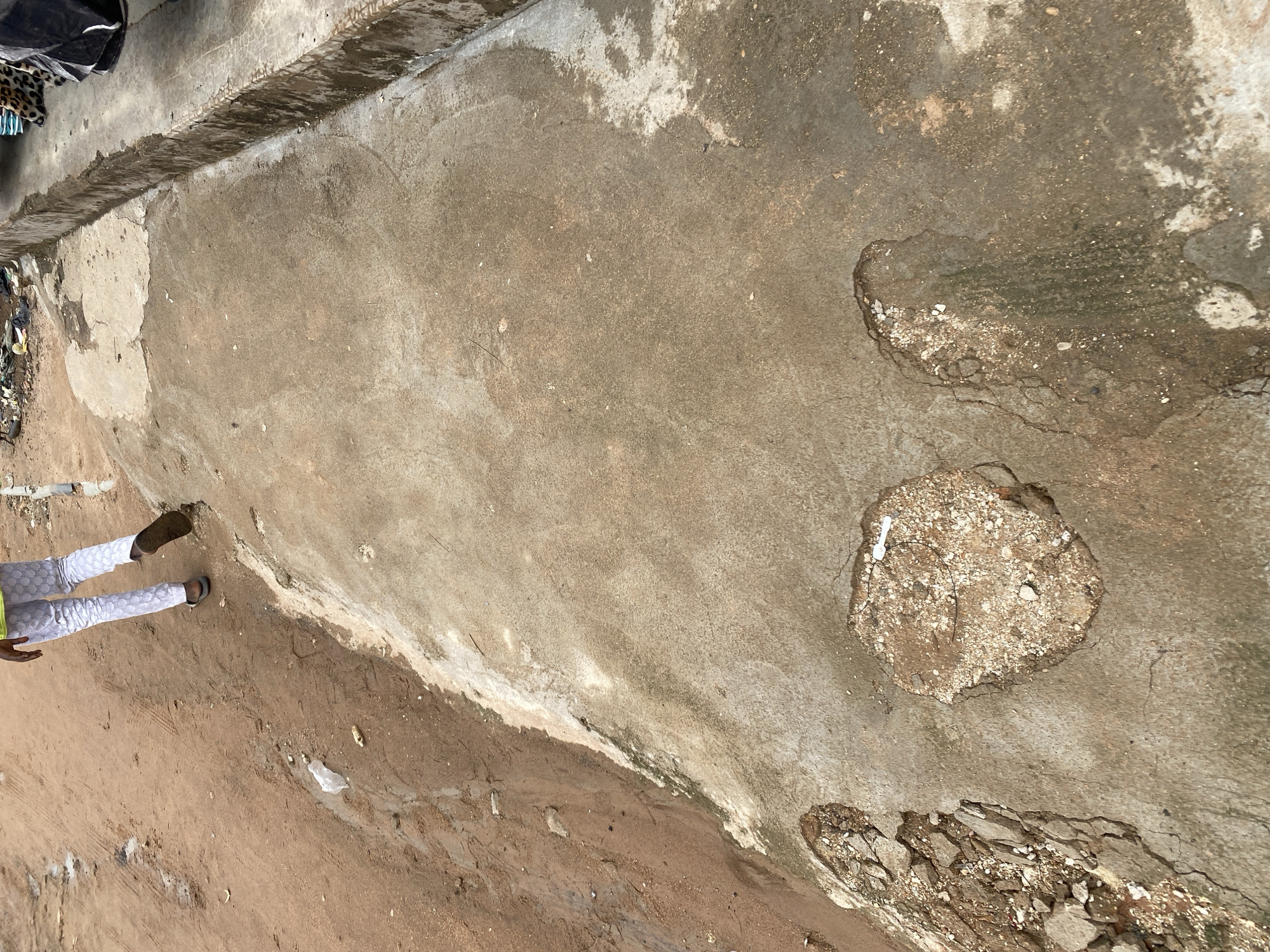 An overhead shot captures a person walking along a dirt and concrete path in Jalingo, Nigeria. The person is wearing white pants with a circular pattern and a lime green shirt. They are seen from the back, walking away from the camera and towards the upper left of the frame, on a dirt path. The path itself is uneven, with patches of loose dirt and some small debris. To the right of the dirt path is a weathered concrete surface. This concrete area is cracked and has several large holes exposing the dirt and gravel beneath. The concrete appears to have water stains and some greenish algae growth in places. Further to the right, a raised concrete curb or step is visible, with debris and possibly some fabric on top of it. The overall scene depicts a somewhat neglected urban environment with evidence of wear and tear on the infrastructure.