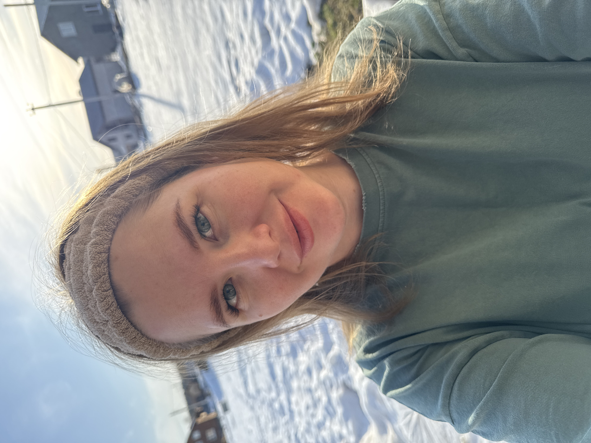 A woman is the primary subject, wearing a tan-colored headband and a green t-shirt. The background shows a snow-covered area with a building visible in the upper left corner. The photograph appears to be taken in Gornyi (Dvurechenskoe S/P), Russia.
