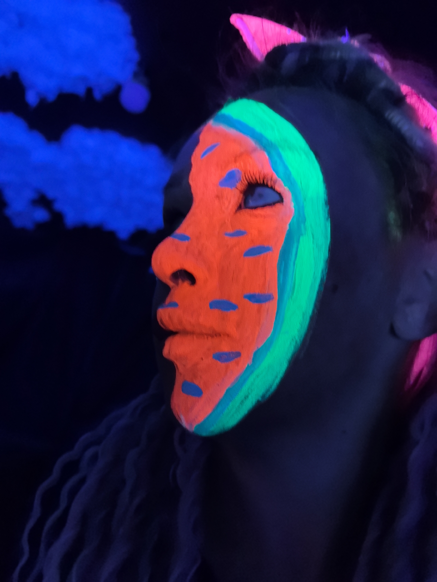 A close-up photograph depicts a person with neon face paint, likely taken under blacklight conditions. The person's face is predominantly covered in orange paint, with blue markings resembling dots or dashes. A stripe of green paint surrounds the orange, highlighting the contours of the face. The background is dark, with some areas illuminated with blue light, possibly representing a cloud-like effect. The subject's hair has sections illuminated with pink light. The specific location in Noviy, Russia, is not discernible from the visual information provided.