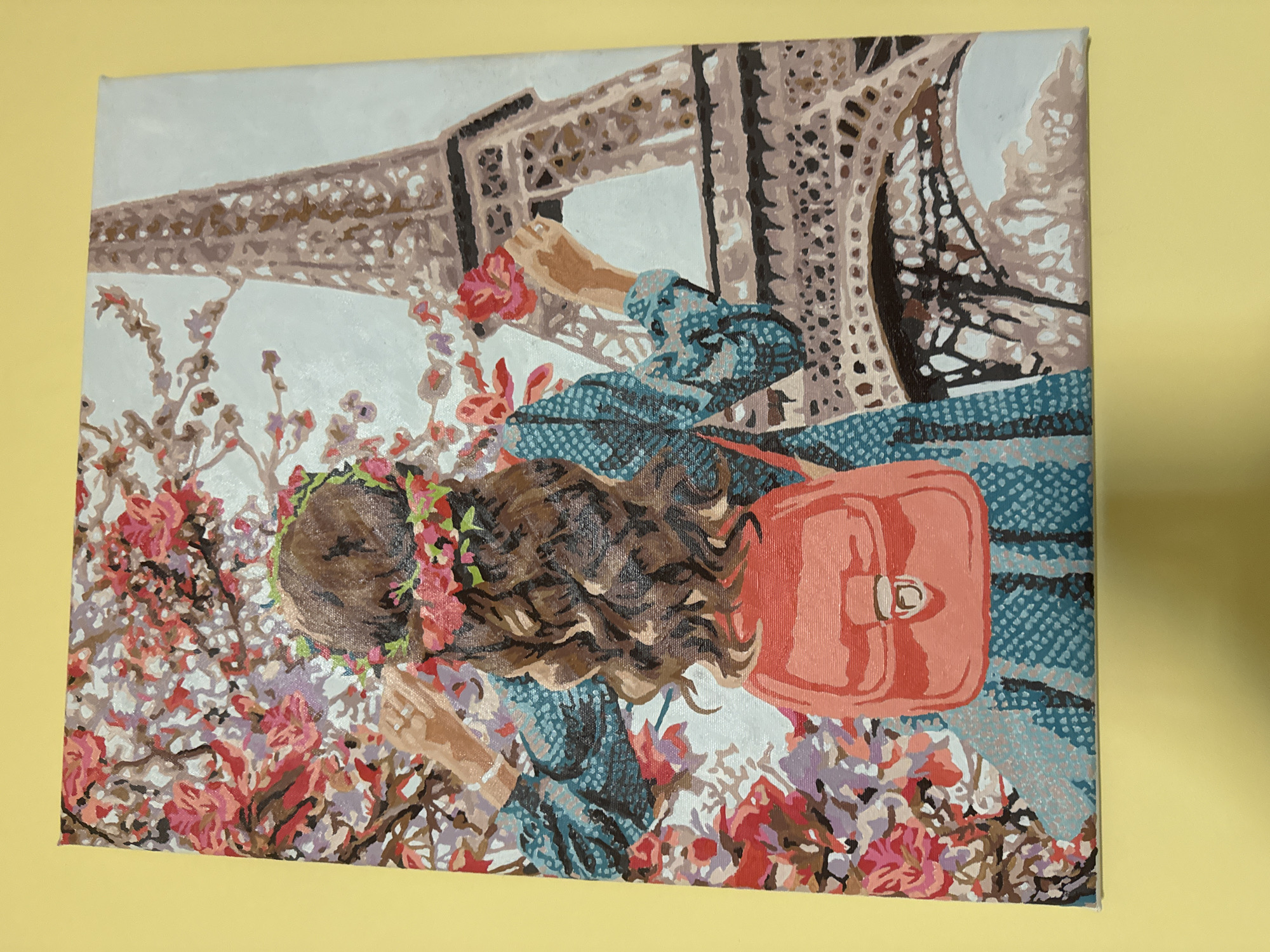 The image shows a painting of a woman with her back turned, standing near the Eiffel Tower in Paris. The woman has brown hair with a flower crown and is wearing a blue dress with white polka dots. She is carrying a salmon-colored purse on her right shoulder. She is holding a red flower in her right hand, reaching towards the Eiffel Tower. In the foreground there are white and pink flowers. The Eiffel Tower is visible in the background. The painting style is reminiscent of paint-by-numbers. The painting hangs against a solid yellow wall.