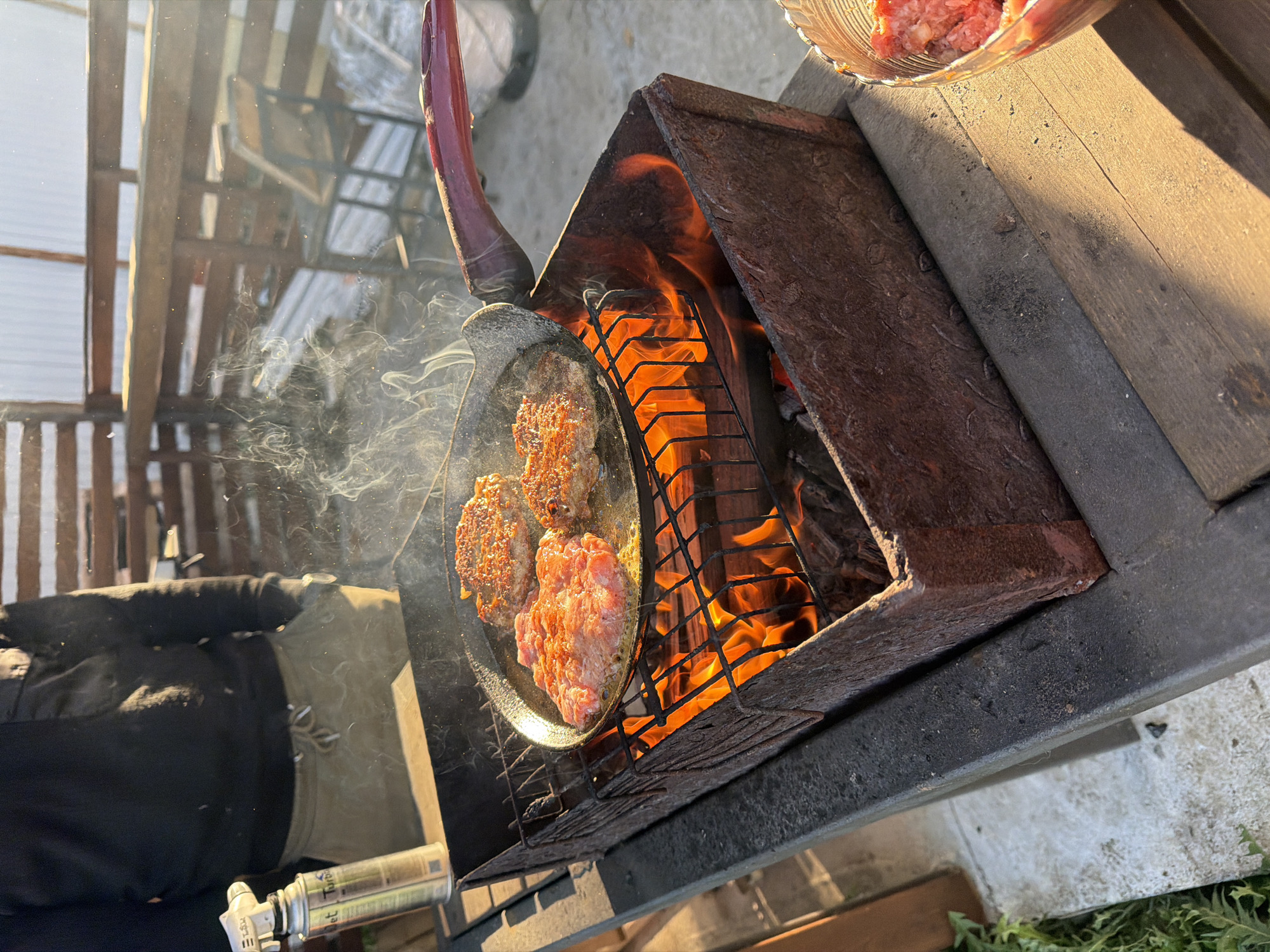 A person is cooking patties over an open fire in what appears to be a backyard or outdoor cooking area. The patties are in a frying pan set over a metal grate above the flames. Smoke is rising from the pan. To the side, there's a bowl of what appears to be raw meat. The fire pit is surrounded by wooden structures. A person wearing a black jacket and beige pants is visible near the grill. A metal canister is on the ground near them. The image shows a sunny, outdoor setting. This scene takes place in the city of Большая Шильна, Russia.