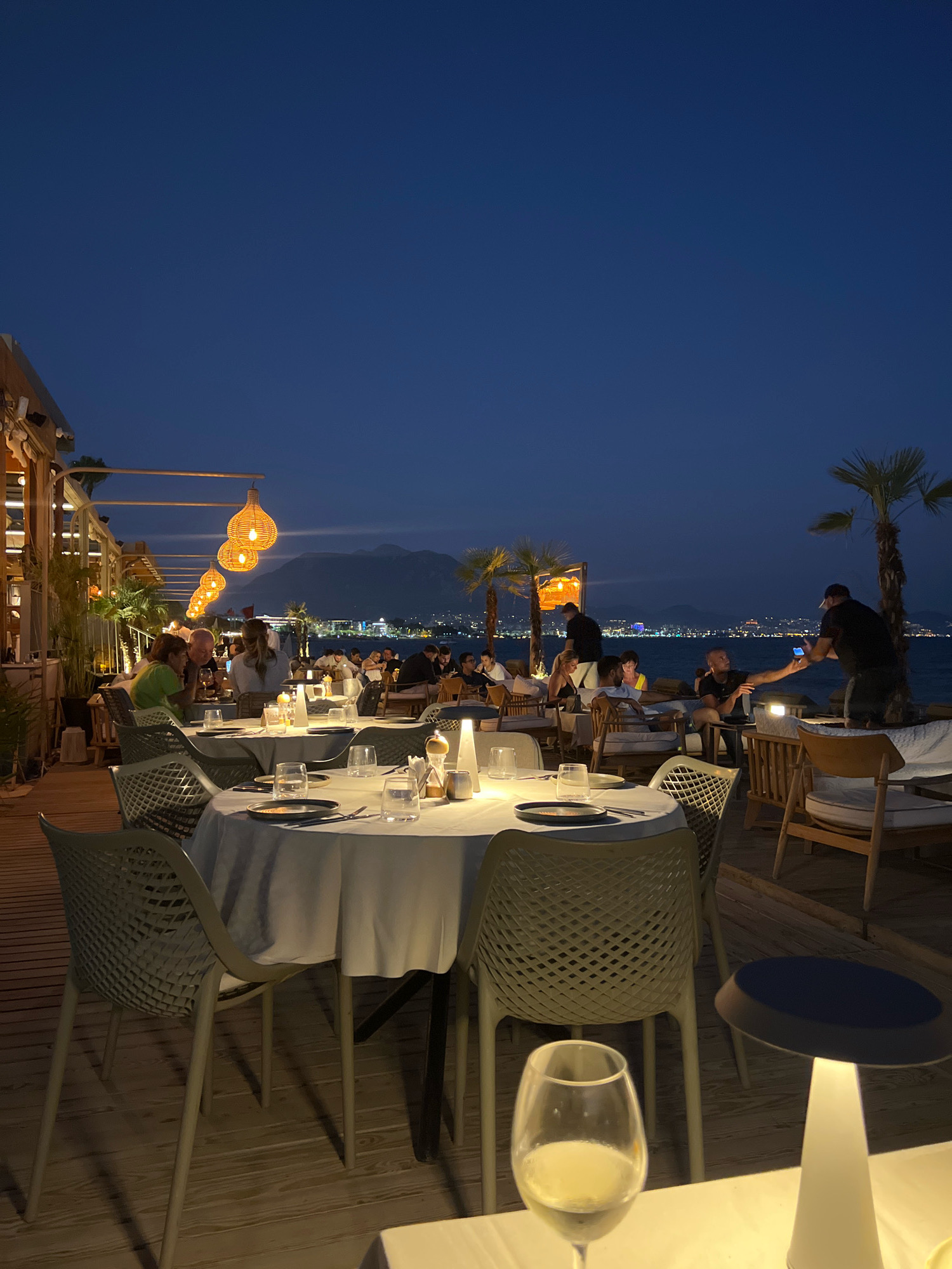 The image depicts a nighttime scene at a beachfront restaurant in Alanya, Turkey. Numerous tables with white tablecloths are set up on a wooden deck overlooking the sea. Palm trees are visible, as are distant city lights along the coastline, suggesting the restaurant is located within or near the city of Alanya. People are seated at the tables, engaged in conversation and dining. The atmosphere is illuminated by hanging pendant lights and table lamps, creating a warm and inviting ambiance. In the foreground, a table is set with glasses and plates, and a glass of white wine is prominently featured. Further back, a man is seen taking a photo with his phone. The sky is a deep blue, characteristic of the evening or night.