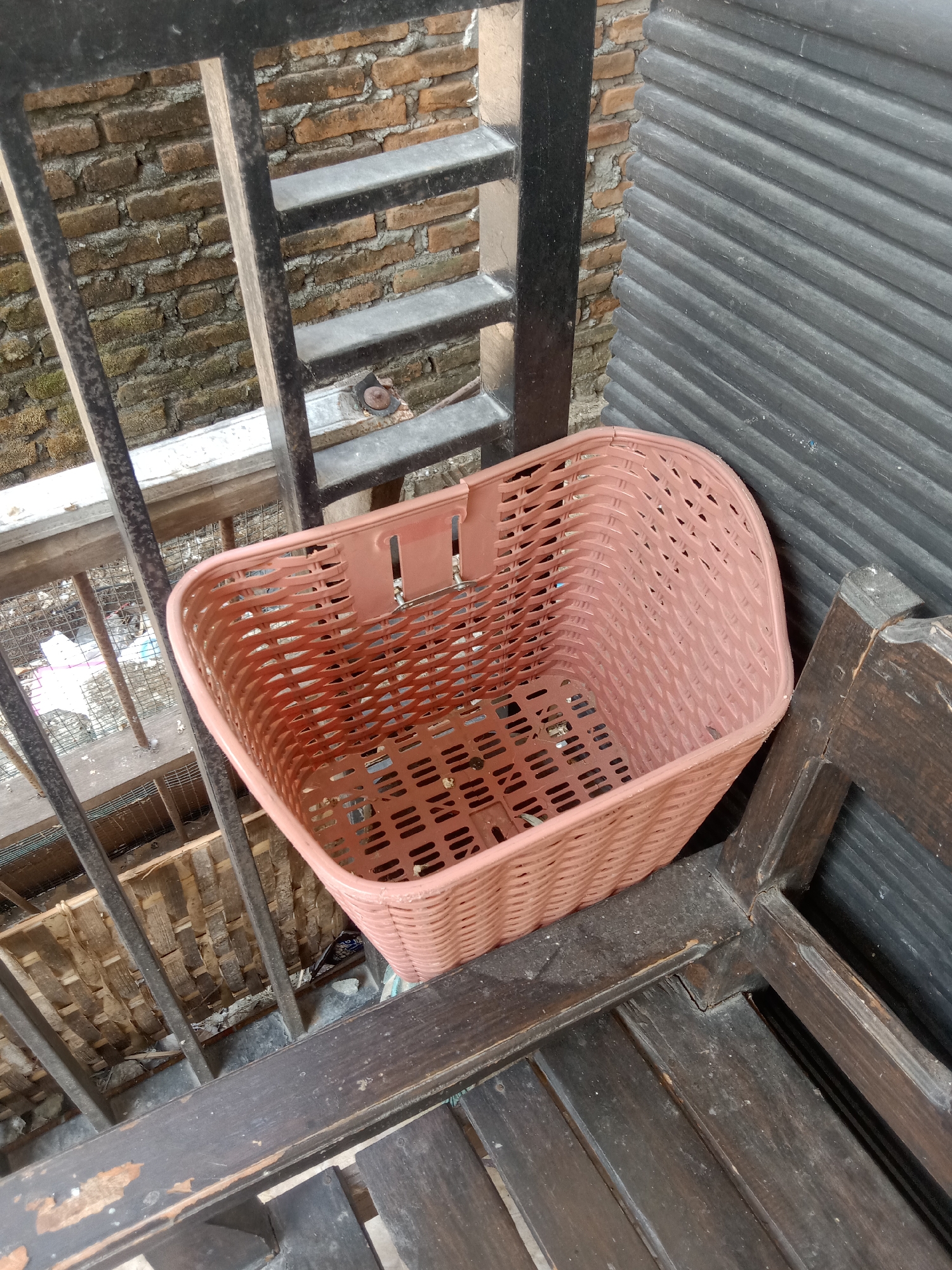 The image shows a pink plastic basket with a woven pattern, placed on a dark wooden bench. The basket is positioned outdoors, with a brick wall in the background and a black metal railing to the left. A dark gray corrugated metal structure is visible behind the basket and to the right. The scene appears to be in an outdoor area, possibly a balcony or a courtyard, with evidence of a slightly neglected environment, including some debris visible below the railing. The specific location in Stiaji, Indonesia, cannot be determined from the visual information alone.