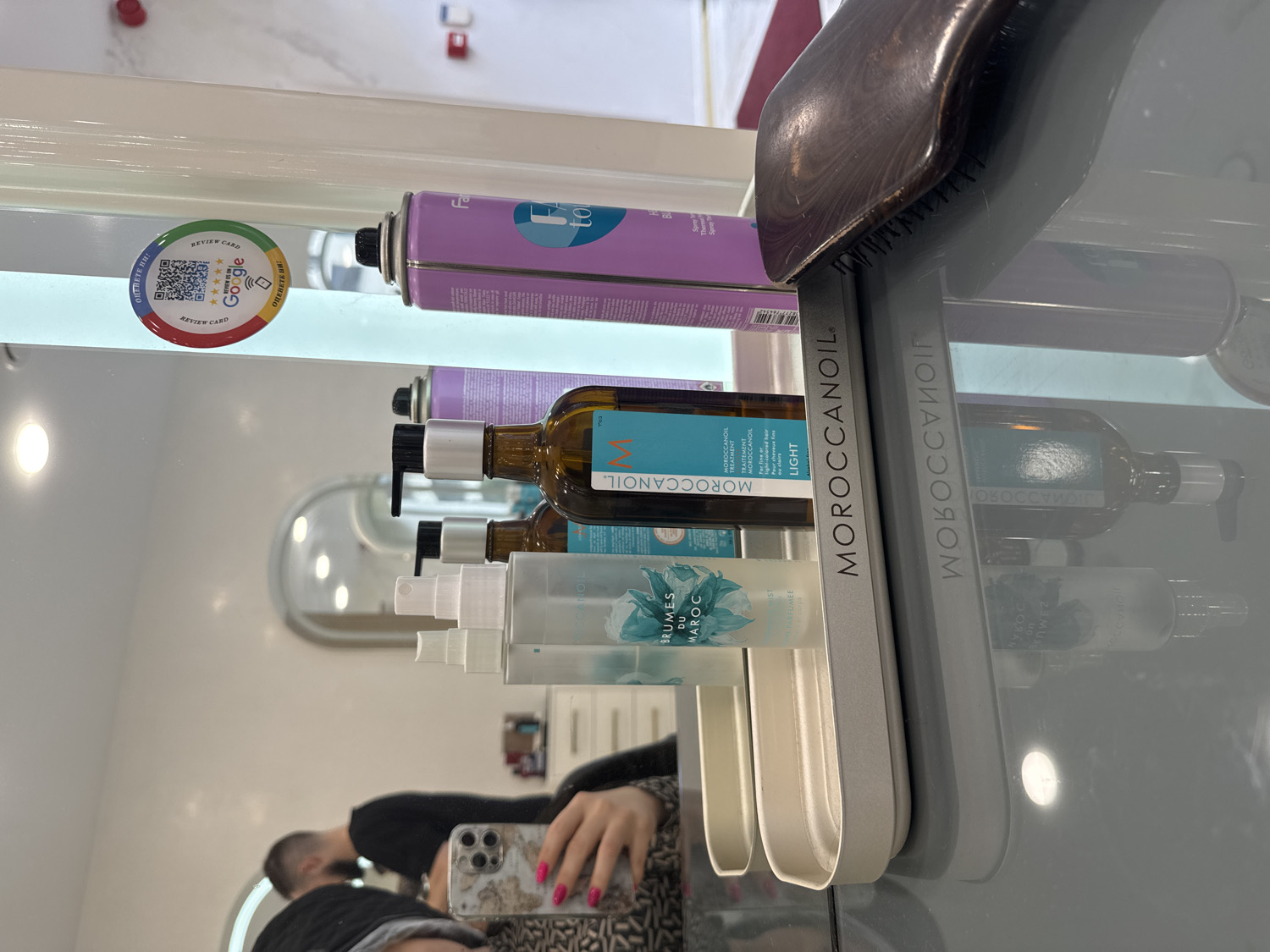 A mirror-reflection shot captures a segment of what appears to be a hair salon or studio in Sofia, Bulgaria. The main subjects are various hair care products displayed on a sleek gray and white stand, reflected in a large mirror.
Prominently featured are several Moroccanoil products: a bottle of Moroccanoil Treatment Light with a pump dispenser, and two clear spray bottles labeled "BRUMES DU MAROC." Above these, a purple aerosol can, possibly hairspray, with "Fa" and "to" visible on its label, is also reflected. A wooden-handled hairbrush rests on the top of the Moroccanoil display stand.
The image is captured by a person whose hand, adorned with pink-patterned nail polish, holds a smartphone with a prominent camera array. Part of the person's reflection, including a bald or closely-shaved head, is visible in the lower left of the frame, indicating they are seated or standing in front of the mirror.
A circular "REVIEW CARD" sticker is adhered to the mirror, featuring a QR code, five stars, the Google logo, and the Bulgarian phrase "ОЦЕНЕТЕ НИ!" (RATE US!), which confirms the location context of Sofia, Bulgaria. The setting is brightly lit, suggesting an indoor environment, likely a professional establishment. The overall impression is one of a modern, well-equipped salon.